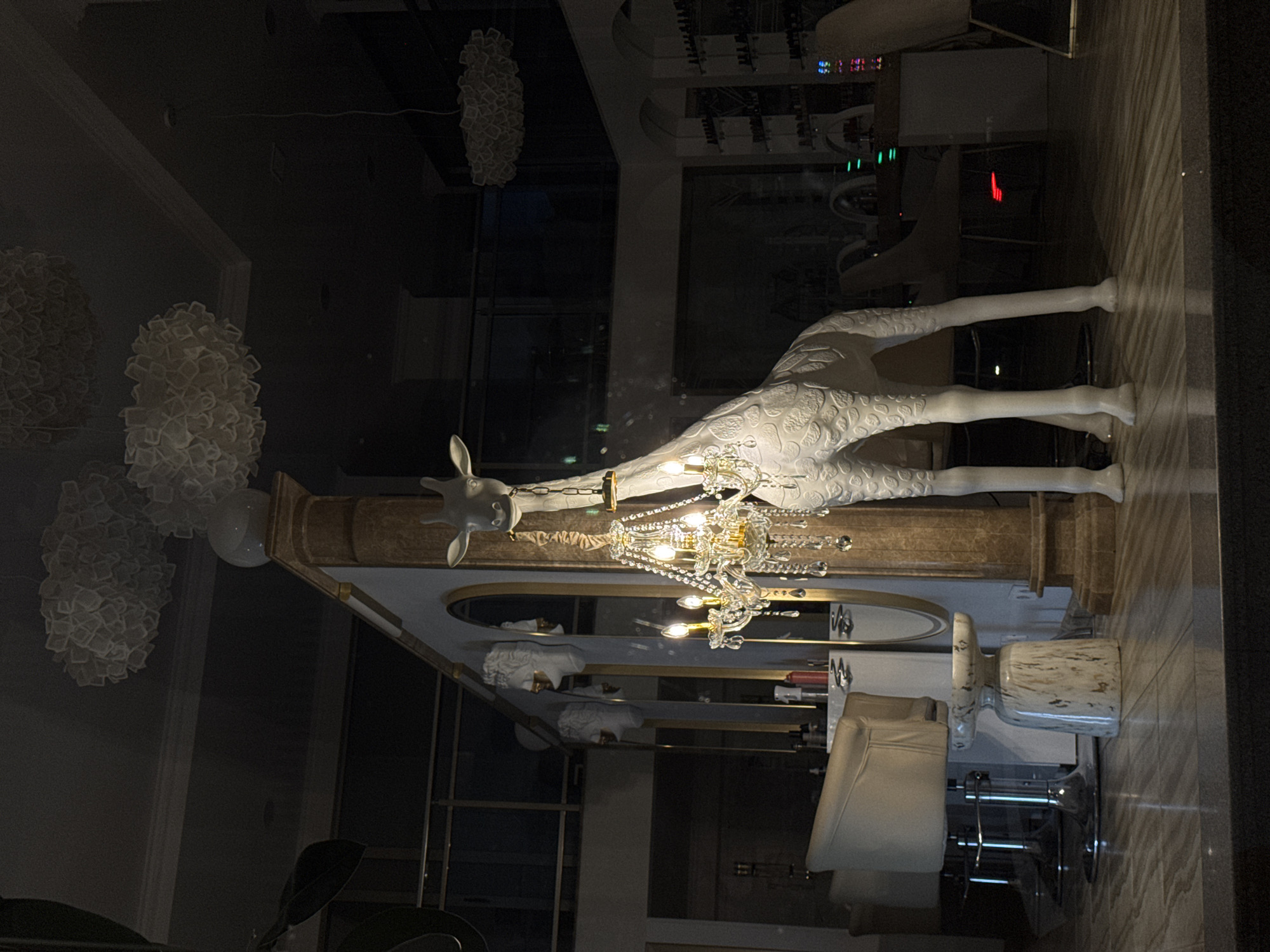 The image depicts an interior scene with a large, white giraffe statue as the central subject. The giraffe is adorned with a crystal chandelier hanging from its neck. The environment appears to be a salon or boutique, with styling chairs, mirrors, and shelves visible in the background. The lighting is dim, with the chandelier and some ambient lights providing illumination. There are no people visible in the image. The location is identified as the city of Batumi, Georgia.