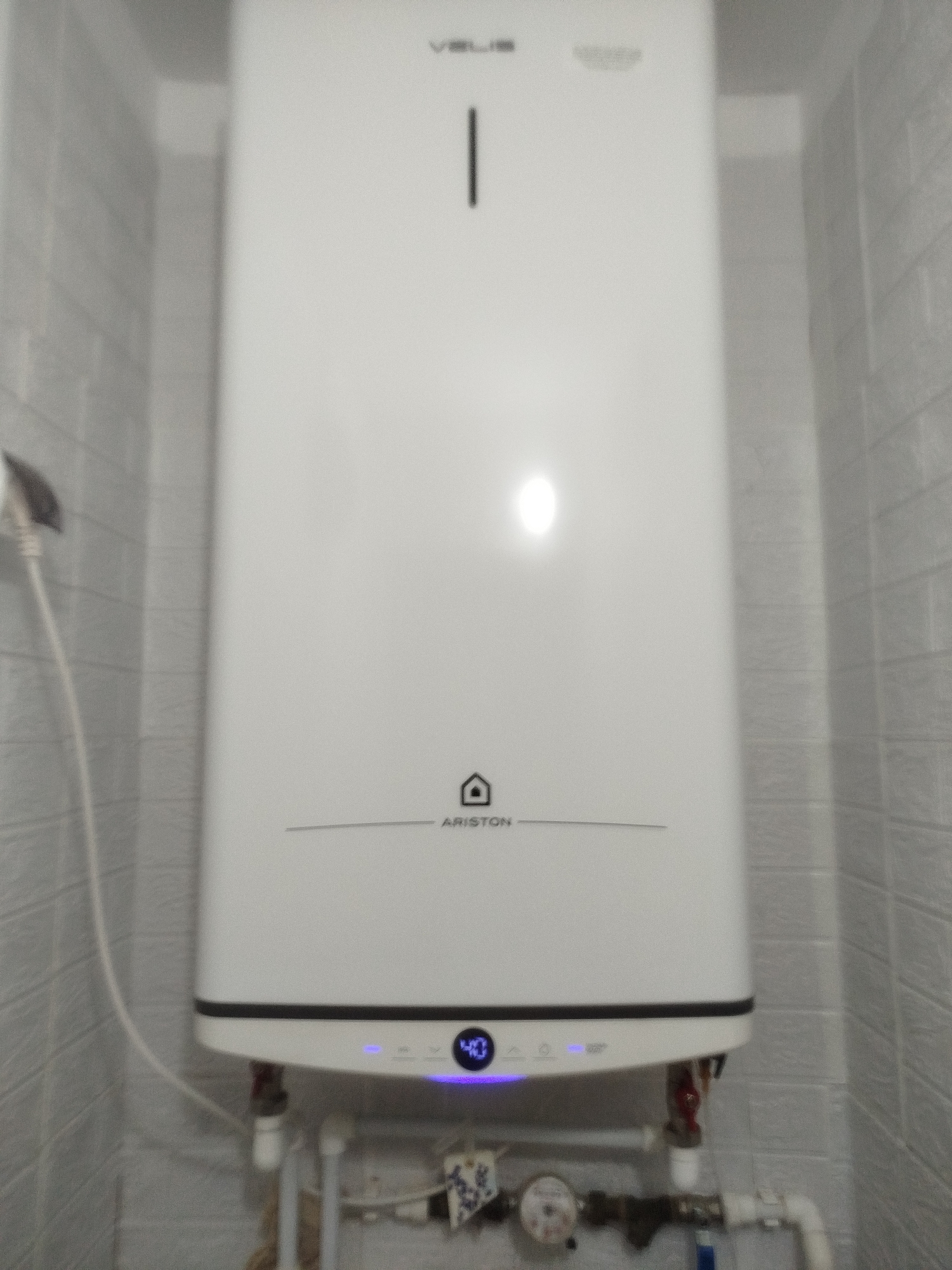 The image shows a white Ariston water heater mounted on a tiled wall. The water heater has a digital display at the bottom showing "40" in blue LED lights, indicating the temperature. Above the display, there's a circular logo with a house icon inside, and the text "ARISTON" below it. The top of the water heater has a vertical black line. To the left of the water heater, a white electrical cord is visible. The tiled wall appears to be in a bathroom or utility room, with plumbing pipes visible at the bottom of the image. The scene is indoors, and the lighting suggests it is daytime or well-lit artificial light. No people are visible in the image.
