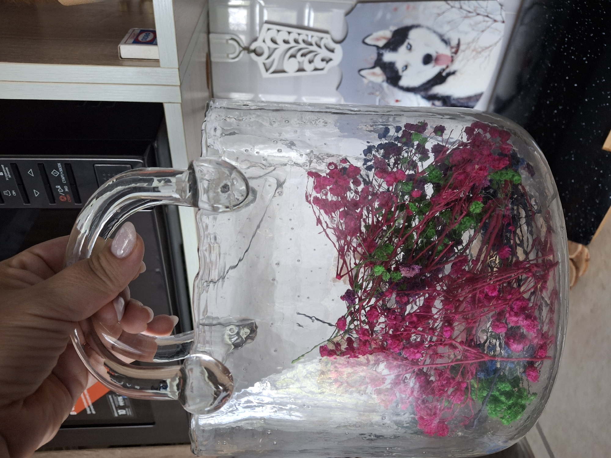 A woman's hand, with painted nails, holds a clear glass bag filled with dried flowers. The flowers are a mix of pink, green, and blue. The bag has two glass handles. In the background, a microwave is visible, along with a shelf containing a small box. A decorative tile with a husky dog image is also present. The setting appears to be indoors, possibly a kitchen.