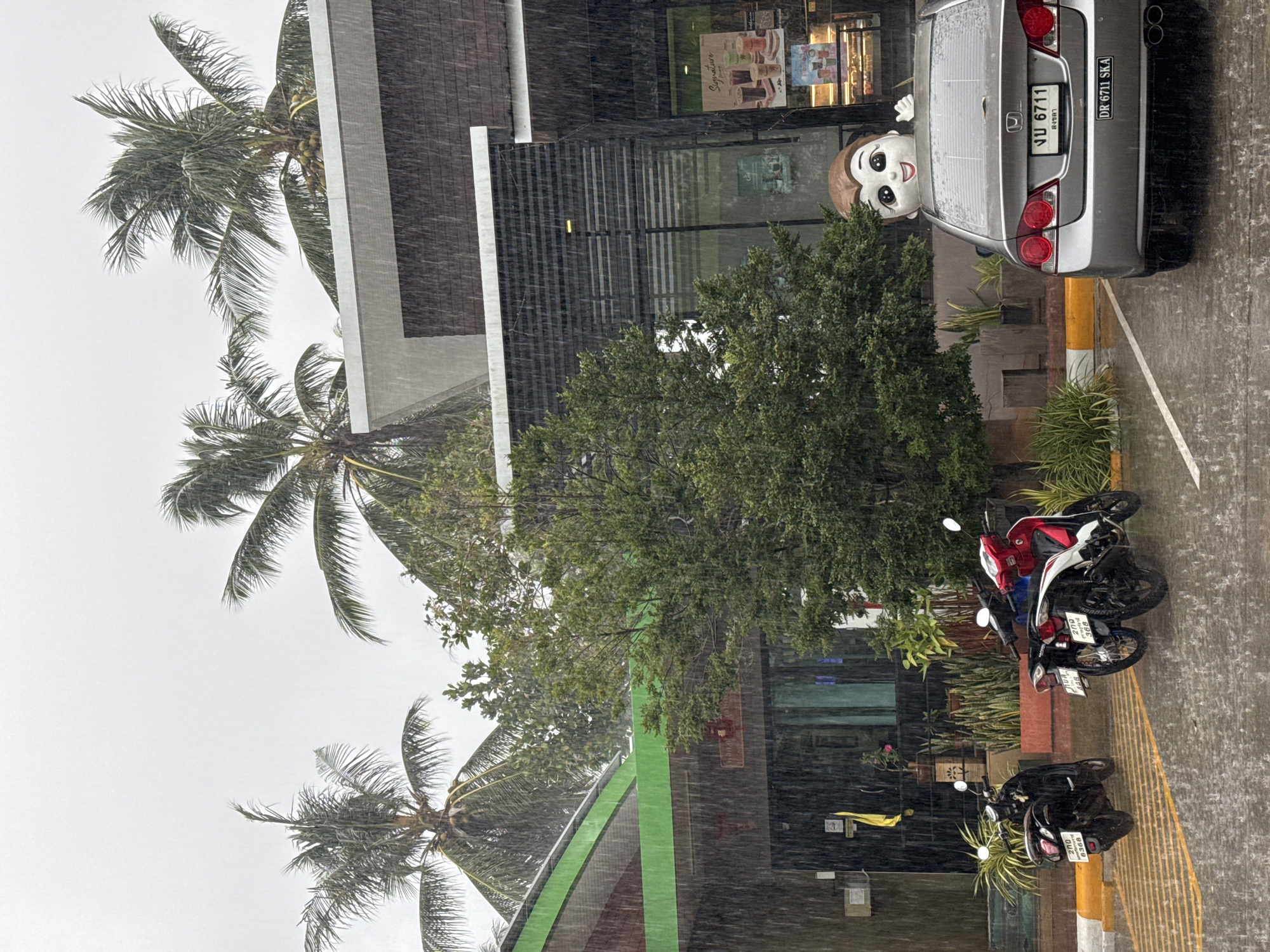 In Ko Samui District, Thailand, a silver sedan is parked in front of a building with a modern, multi-story design. Heavy rain is falling, with visible streaks of water against a gray sky and obscuring some details of the environment. A large, cartoonish figure of a boy's head with wide eyes and a smiling mouth is peeking over the trunk of the sedan. Two motorcycles are parked to the left of the sedan. The building appears to be a commercial establishment, possibly a cafe or shop, with windows showcasing displays and posters. Palm trees are visible in the background, their fronds glistening with rain. The ground is wet and reflects the light.