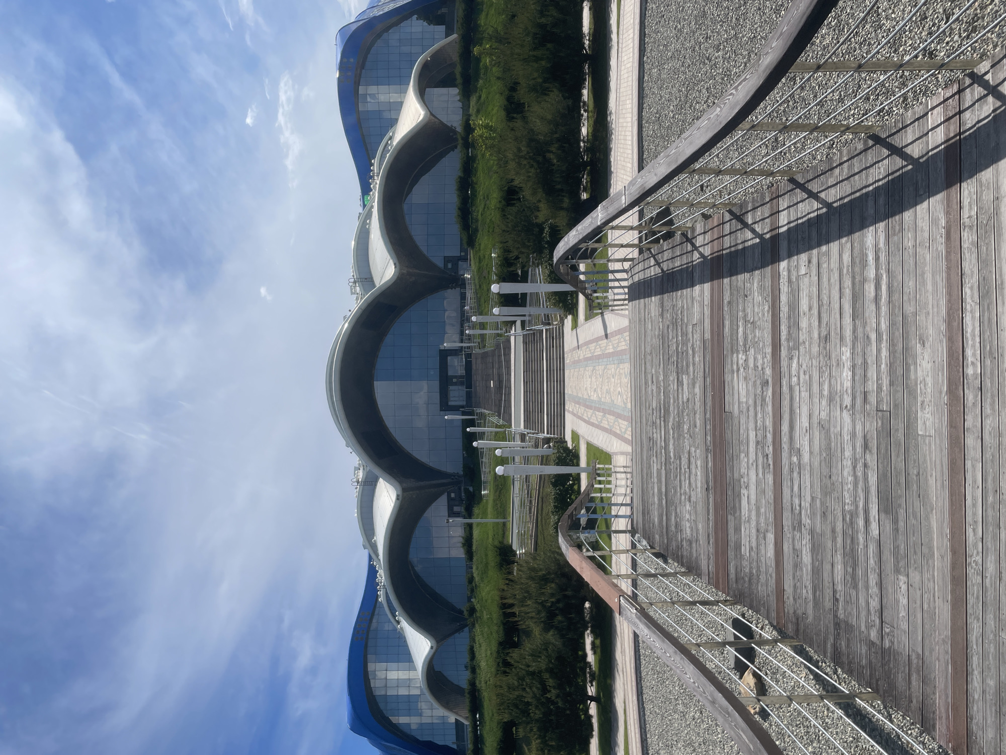 The image shows the exterior of the Primorye Aquarium located in Russky, Russia. A wooden walkway with metal railings leads towards a grand entrance with wide stairs. The building itself features a distinctive modern architectural design with blue curved roofs and large glass windows. Lush green bushes and trees surround the structure, and the sky above is partly cloudy with a bright blue hue. The overall scene depicts a clear day with ample sunlight casting shadows on the walkway.