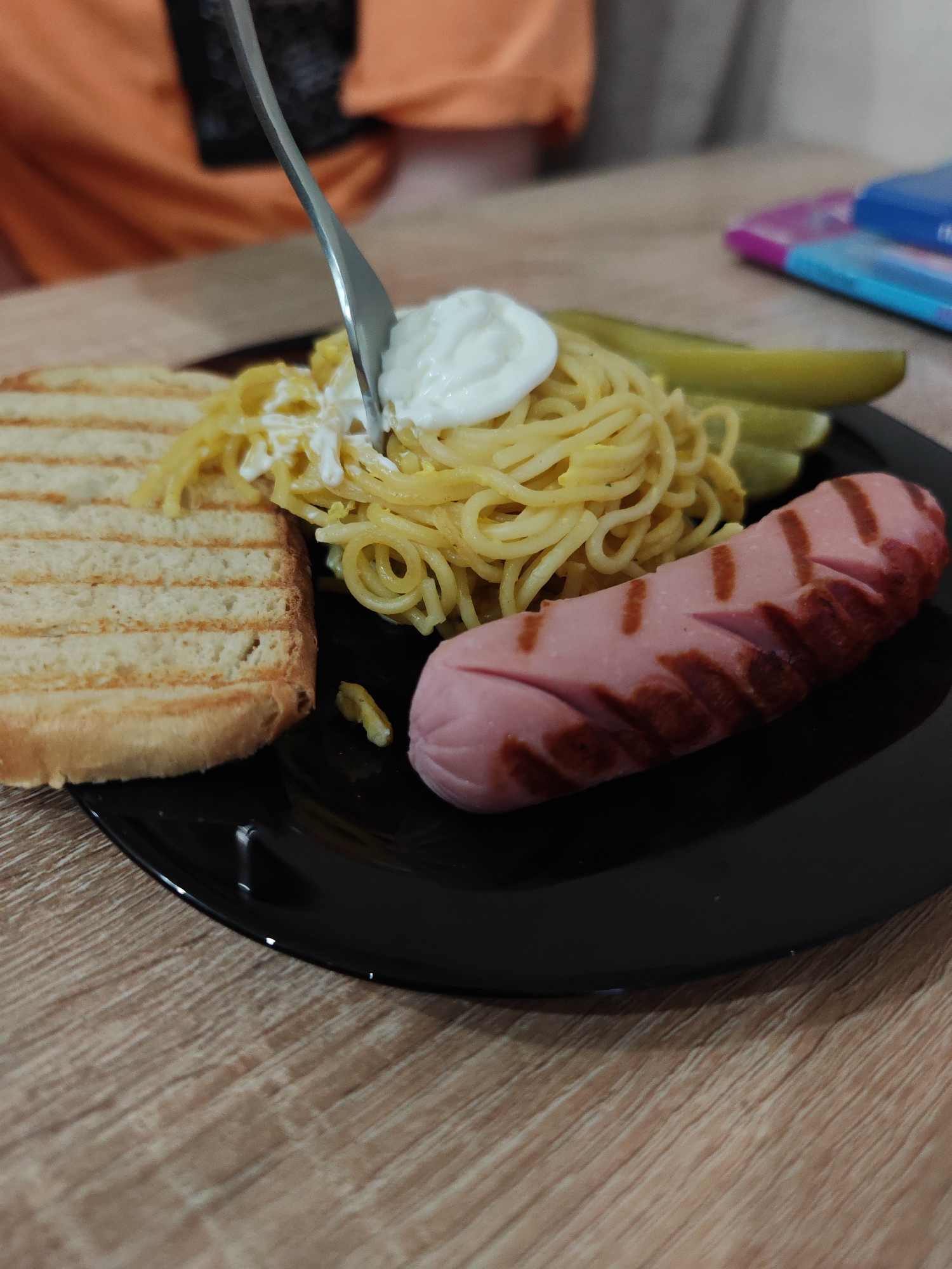 The image shows a close-up of a meal served on a wooden table in Hrodna, Belarus. On a black plate, a portion of spaghetti, possibly mixed with scrambled eggs, is visible. A dollop of a white creamy sauce, likely sour cream or mayonnaise, rests on top of the spaghetti, with a silver fork actively plunged into it, suggesting someone is eating.
To the right of the spaghetti, a grilled sausage or hot dog is presented, featuring distinct dark grill marks and diagonal cuts on its surface. Two green pickled cucumbers are placed beside the pasta and sausage. To the left, a thick slice of toasted bread with prominent grill lines occupies part of the plate.
In the blurred background, a person wearing an orange shirt is partially visible, along with some colorful objects that appear to be stacked notebooks or books. The table surface is light-colored wood with a visible grain. The scene depicts a casual indoor meal. There is no visible text in the image.