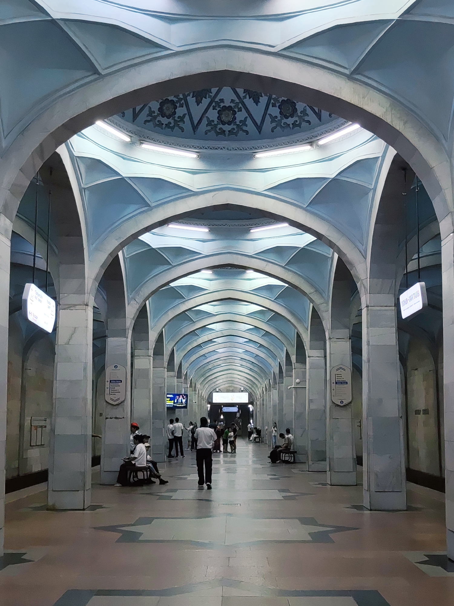 The image depicts the interior of a subway station in Tashkent, Uzbekistan, specifically likely at the Mustaqillik Maydoni (Independence Square) station, given its distinctive architectural style. The station features a grand, symmetrical design with a long, vaulted corridor lined with arches and supported by massive stone pillars. The ceiling is a prominent element, decorated with a light blue geometric pattern and intricate floral motifs near the center. Fluorescent lighting is integrated into the ceiling structure, illuminating the space. The floor is paved with large tiles in varying shades of pink and gray, with a distinctive geometric pattern. Several people are visible in the scene, some walking, others sitting on benches, and a few standing around. The individuals are casually dressed, suggesting an ordinary day in the public transportation system. Signage is visible on some pillars and hanging from the ceiling, indicating station names or directions. The overall atmosphere is one of spaciousness and architectural grandeur.