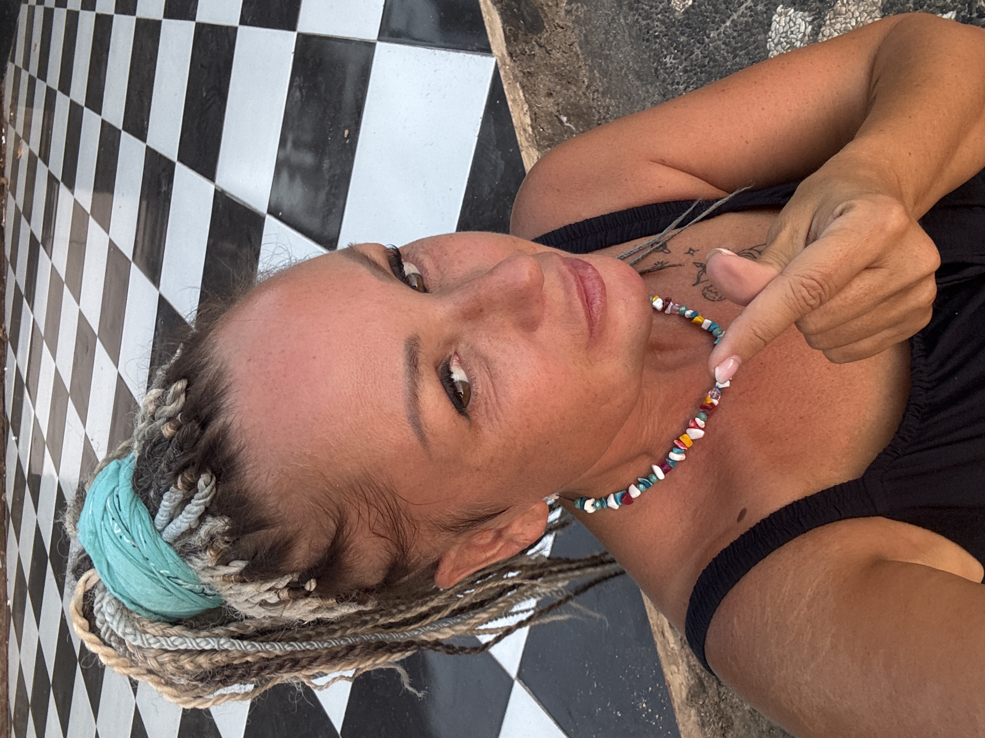 A medium shot image shows a woman in the city of Kuta, Indonesia. She is in the foreground, facing the camera with her left shoulder forward. She has long braided hair of various colors, including blonde, grey, and brown, tied up with a turquoise bandana. She is wearing a black tank top and a colorful beaded necklace. Her right hand is raised, and her thumb and index finger are extended, forming a heart shape gesture. She has freckles on her face and chest. Behind her, a black and white checkered floor is visible. The lighting suggests it is daytime.