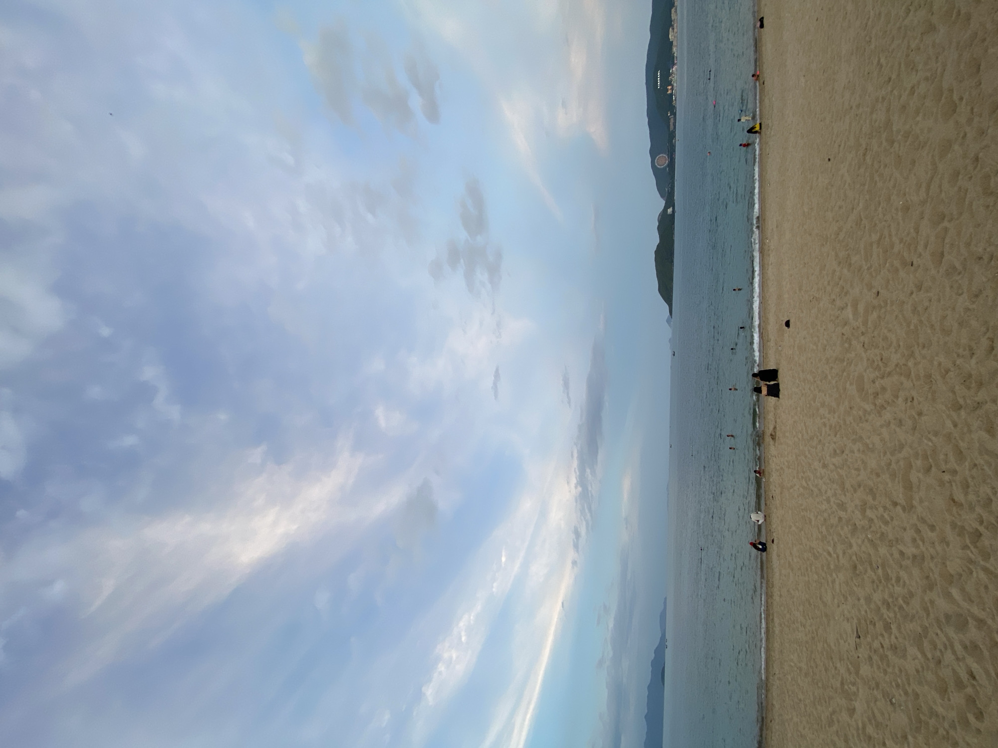 The image depicts a tranquil beach scene in Nha Trang, Vietnam. The foreground is dominated by a wide expanse of sandy beach with subtle footprints visible. In the mid-ground, gentle waves lap the shore, and several individuals are present in the water, some swimming and others wading. Along the waterline, a few people are seated or standing on the sand.
The background features a calm sea stretching towards a horizon marked by distant, verdant islands. The sky above is a canvas of soft blues and whites, with wispy clouds streaking across it, suggesting either early morning or late afternoon light. On the far right of the horizon, a large, circular illuminated object is visible atop a dark, hilly landmass, possibly a landmark or part of a resort. The overall atmosphere is one of peaceful recreation and natural beauty.