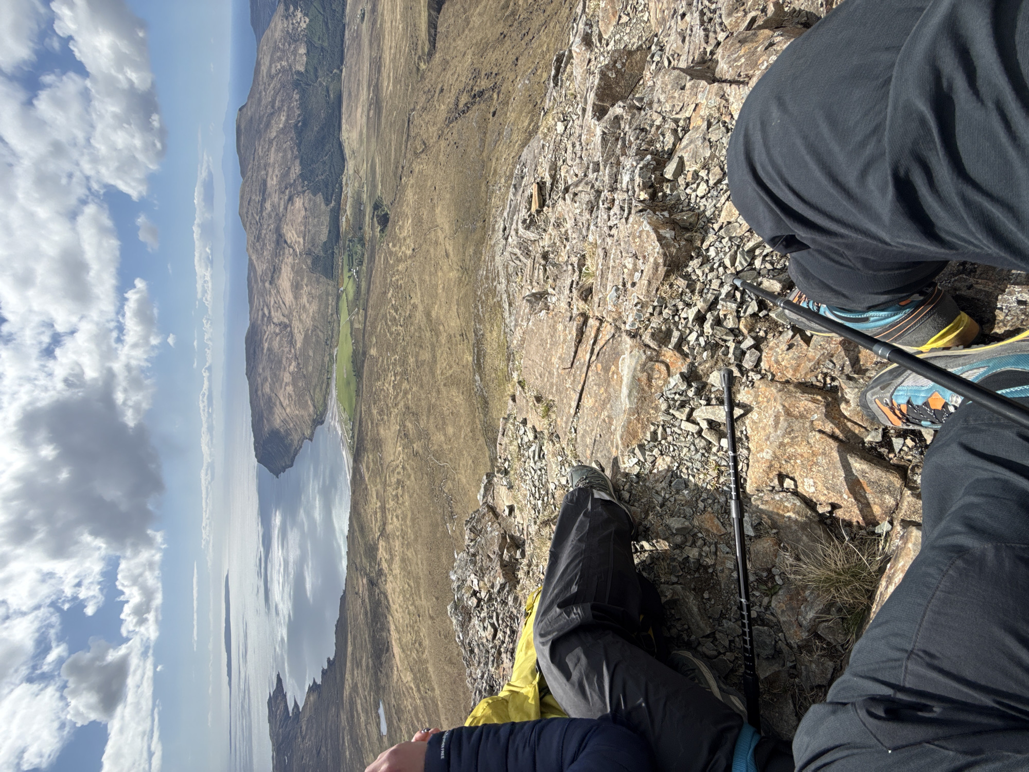 A high-angle, outdoor shot shows two people sitting on a rocky incline, with a sweeping view of a landscape including a body of water, grassy terrain, and distant hills, under a sky with scattered clouds. The people are partially visible from the mid-thigh down, wearing dark trousers and hiking shoes. One person has hiking poles placed near them. The landscape stretches out horizontally, showing a large body of water reflecting the sky, next to rolling grassy hills leading to distant mountains. The sky is partly cloudy. The exact location and city are undetermined.