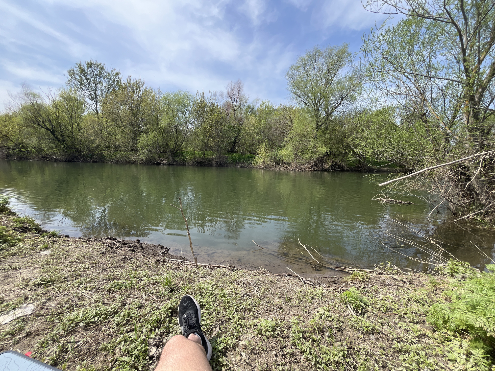 The image depicts a body of water, possibly a river or a wide stream, surrounded by vegetation. The water is calm, with a slightly greenish hue. The banks are covered in grass and small plants. On the right side of the frame, branches extend from the edge of the water. In the foreground, there is a view of a person's leg and foot wearing a black athletic shoe. The location is Unknown, Unknown.