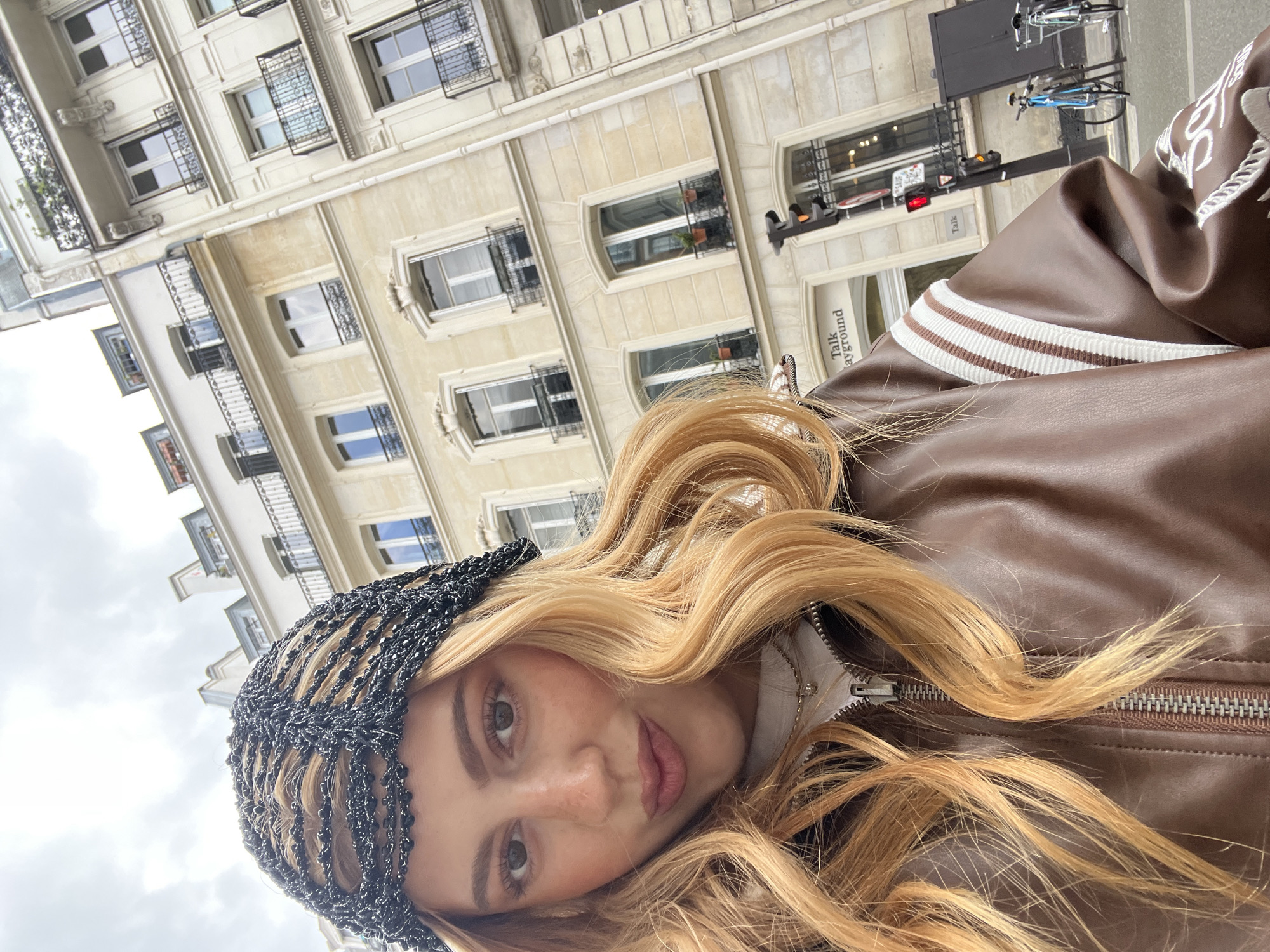 A woman with blonde hair, wearing a dark beaded headband and a brown leather jacket with white stripes on the sleeve, is positioned in front of a Parisian building. The building has multiple stories with windows, some of which have wrought iron decorations. A sign that says "Talk ayground" is visible near one of the entrances. A bicycle is parked behind a traffic light in the background. The sky is overcast.