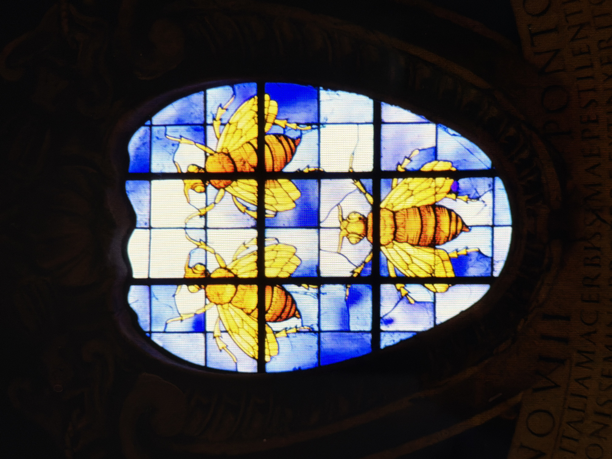The image shows a stained-glass window with three yellow bees against a blue and white checkered background. The window is oval-shaped and set within a dark, ornate frame. Below the window, a stone inscription with Latin text is visible. This stained-glass window is located in the National Academy of Arts of Ukraine in Kyiv, Ukraine.