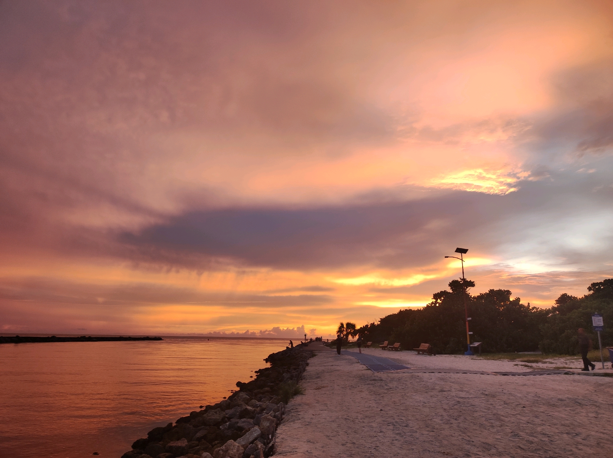 At sunset, a wide, calm body of water reflecting the orange and pink hues of the sky stretches across the left of the frame. A rocky embankment lines the edge of the water, creating a man-made barrier. A paved walkway runs parallel to the embankment, with several people scattered along it, some appearing to be fishing or walking. To the right of the walkway, a sandy area is dotted with park benches and lush green trees. A solar-powered lamppost stands tall amidst the trees. A person in the foreground is walking on the sandy path. The sky is filled with dramatic clouds painted in shades of orange, pink, and purple, indicating a beautiful sunset over the coastal area of Nokomis, Florida.