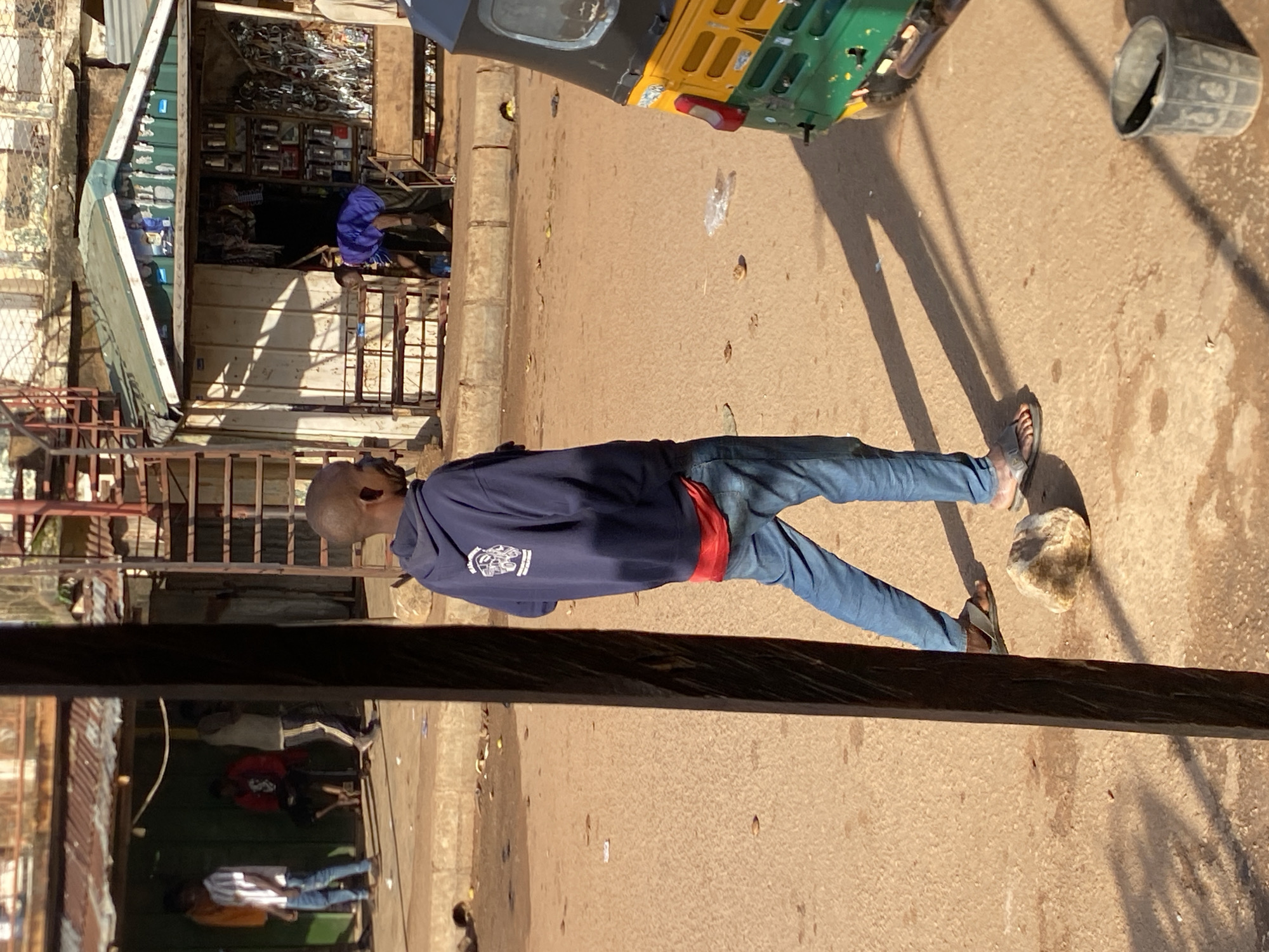 The image, correctly oriented, captures a sunlit street scene in Bukuru, Nigeria. In the foreground, a man with short hair stands with his back mostly to the viewer, facing left. He is dressed in a dark blue hooded sweatshirt featuring a white, somewhat indistinct logo or emblem on the back, blue jeans, and grey sandals. A red band or fabric is visible around his waist. A light-colored rock is situated on the reddish-brown, unpaved ground near his right foot.
Long, distinct shadows stretch across the street from the man and other objects, indicating bright sunlight, likely mid-morning or late afternoon. To the upper right, a yellow and green three-wheeled auto-rickshaw (tuk-tuk) is parked or moving. A grey plastic bucket lies on its side further to the right.
Behind the man, a small shop or stall with a green and white awning is open for business. Its interior, framed by metal railings, reveals shelves packed with various items, possibly general merchandise or auto parts. A person in blue attire is partially visible inside the shop.
The lower section of the image shows a dark, reflective surface, possibly a window or a vehicle, reflecting parts of the street and several individuals. These reflections include a person in a white top and blue jeans, another in a red top, and a third in a white shirt with striped pants, suggesting ongoing street activity. The overall setting is a bustling commercial or market area, consistent with an urban or semi-urban environment in Nigeria. No distinct text is legible on any object.