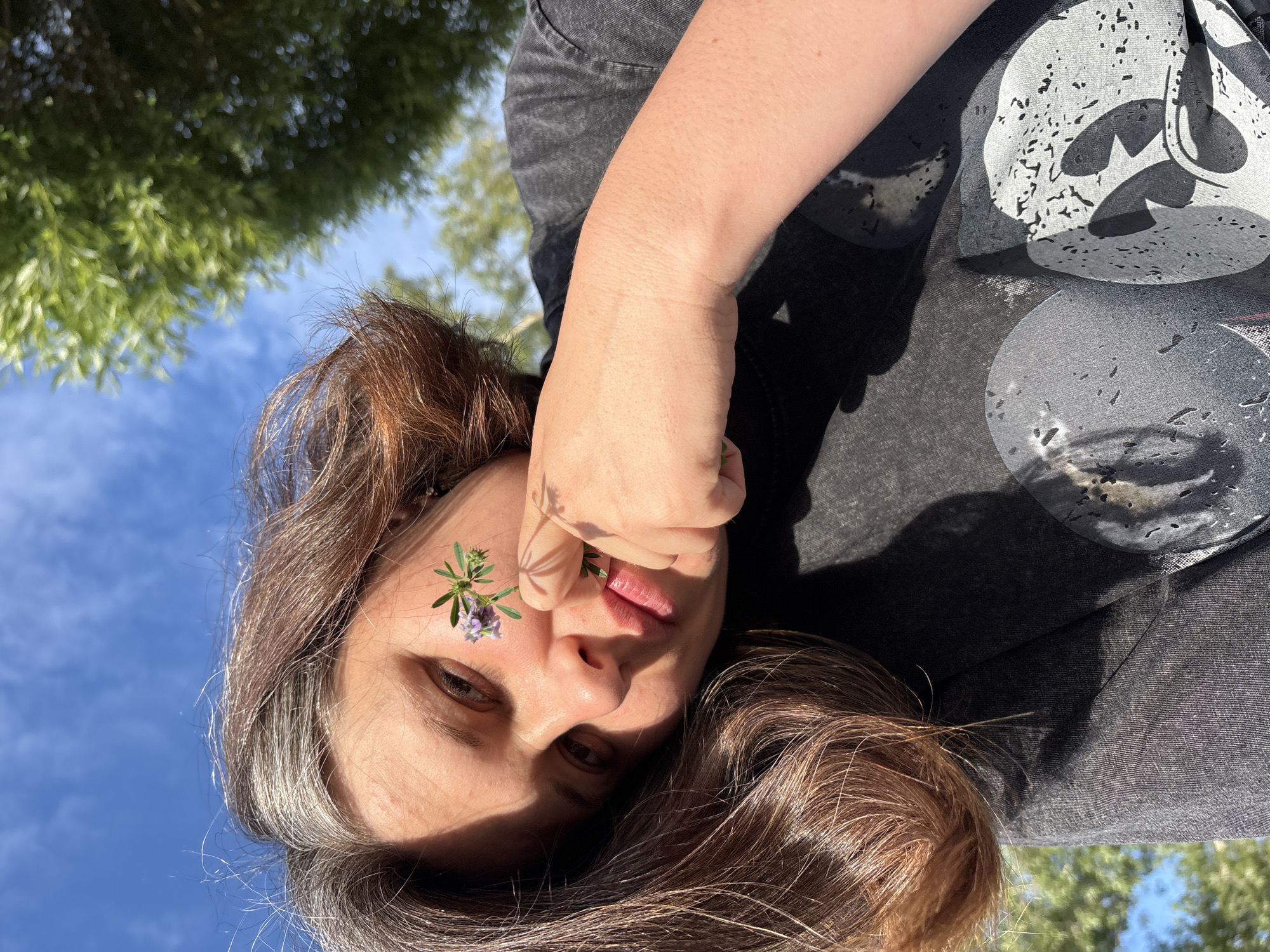The media file shows a close-up, low-angle shot of a person lying down outdoors, looking up towards the sky. The person has long, brown hair with some gray streaks. A small sprig of purple flowers is placed on their right cheek, near the eye. Their face is partially obscured by their right hand, which is held up near their mouth.
The background consists of a bright blue sky with scattered white clouds, and the tops of green trees are visible. The lighting suggests it is daytime, likely late morning or early afternoon, with clear weather. The person is wearing a dark t-shirt with a graphic print. The overall impression is one of casual, relaxed outdoor activity, possibly in a park or natural setting. No text is visible in the media file.