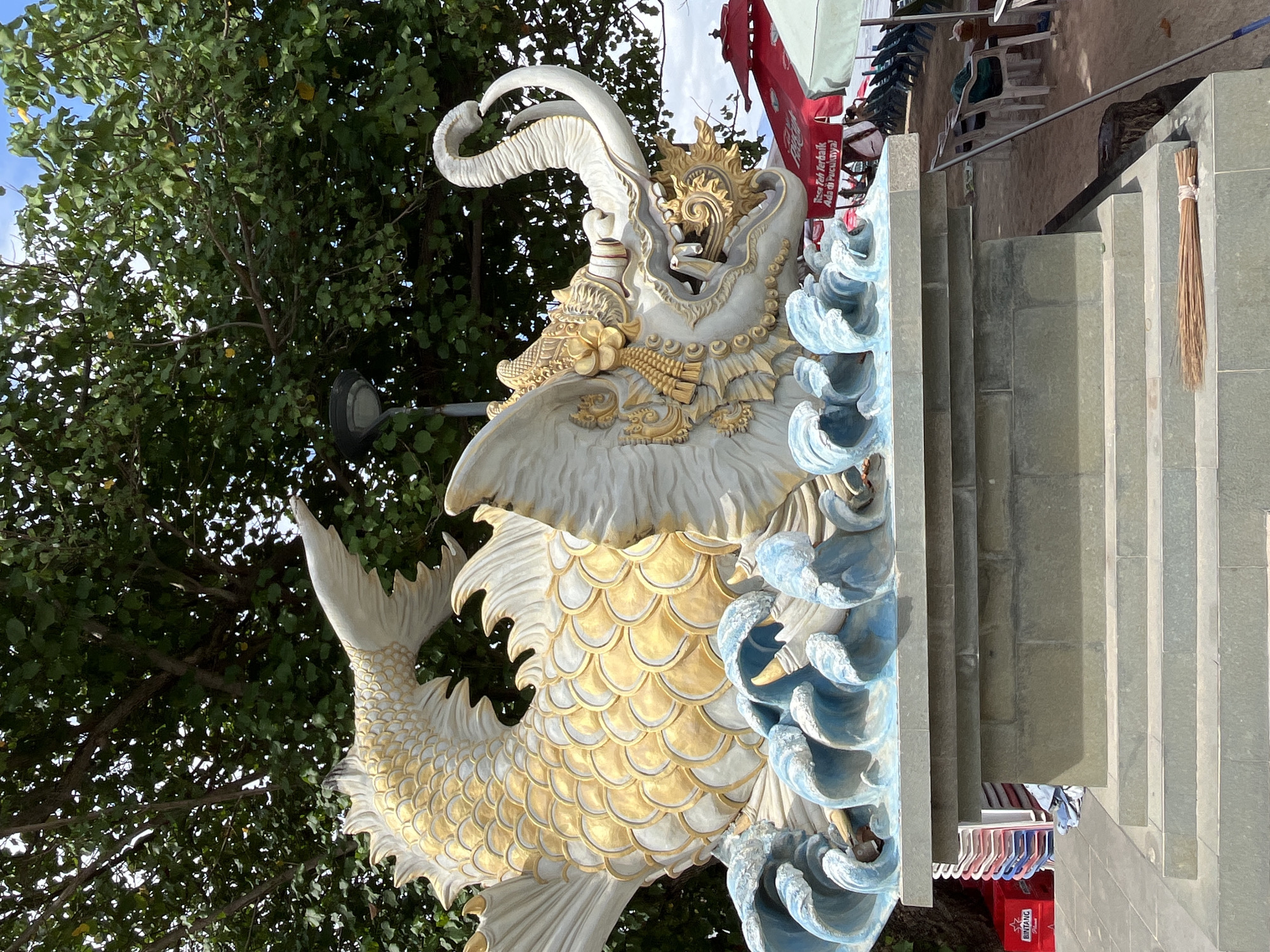 This media file captures a striking close-up of a large, ornate statue, likely a mythical creature resembling a dragon or a koi fish, positioned outdoors. The statue is predominantly white with accents of gold on its scales, headdress, and decorative elements. It is rendered with intricate detail, featuring a detailed head with horns or antennae, a finely scaled body, and a flowing tail. The statue is depicted amidst stylized waves, colored in shades of blue and white, suggesting a marine or aquatic theme.
The background reveals a lush green tree with dappled sunlight filtering through its leaves, indicating a daytime setting with clear weather. To the right of the statue, a set of stone stairs ascends, with a metal railing. At the top of the stairs, several beach chairs are visible, along with red and white umbrellas, hinting at a beach or resort environment. A straw broom, tied with white string, is propped against the stone wall beside the stairs.
Below the stairs, a red crate with the word "BINTANG" printed on it is partially visible, along with a rack of colorful clothing or towels, possibly for sale or rent at a beachside establishment. A small red sign with white text is attached to one of the umbrellas, but the text is difficult to decipher clearly. The overall impression is of a vibrant, culturally rich location, likely Kuta, Indonesia, given the statue's style and the beach setting. No people are clearly visible in the frame.