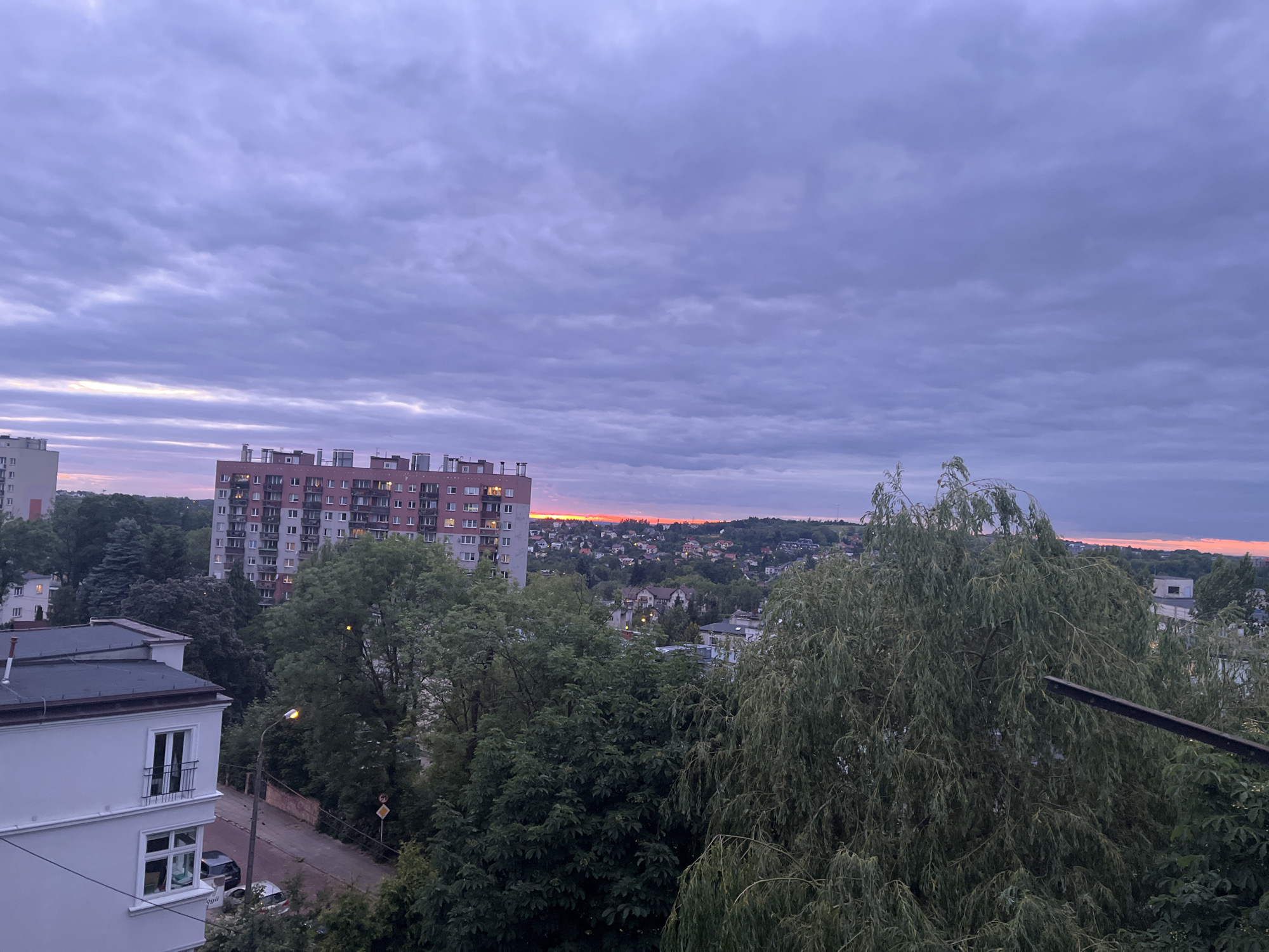 A medium-shot image, taken from a high vantage point, captures a city skyline at dusk. The sky is a canvas of muted purples and grays, with a faint band of orange light on the horizon. Dominating the middle ground is a tall, red brick apartment building with numerous windows and balconies. In the foreground, lush green trees partially obscure the view, with a prominent weeping willow on the right side of the frame. The lower left features the white facade of a residential building with windows and a balcony. A street with parked cars and a lamppost can be seen between the buildings. The overall scene depicts a quiet urban setting as day transitions into night.