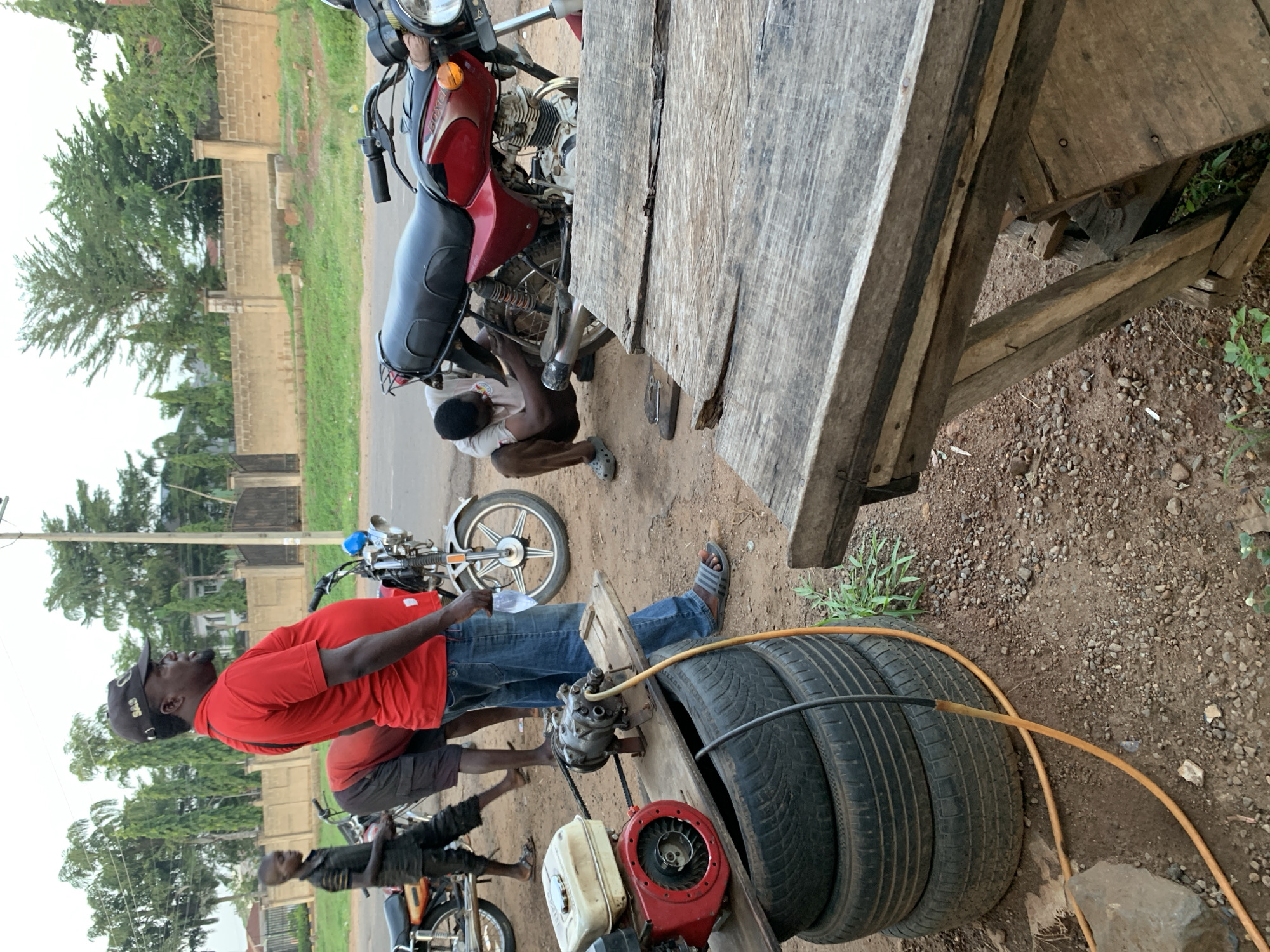 The image shows an outdoor scene in what appears to be a motorcycle repair shop in Gbeleko, Nigeria. In the foreground, a man wearing a red shirt, blue jeans, a black baseball cap, and sandals stands next to a small motor, tires, and a wooden platform. An orange hose snakes across the ground near the tires. To the left, a second man in a dark shirt and shorts is working on a motorcycle. Behind him, another motorcycle is parked. A third man is also working on a motorcycle beneath a large wooden table. In the background, there are trees, power lines, and a fence bordering a road. The ground is dirt and gravel.