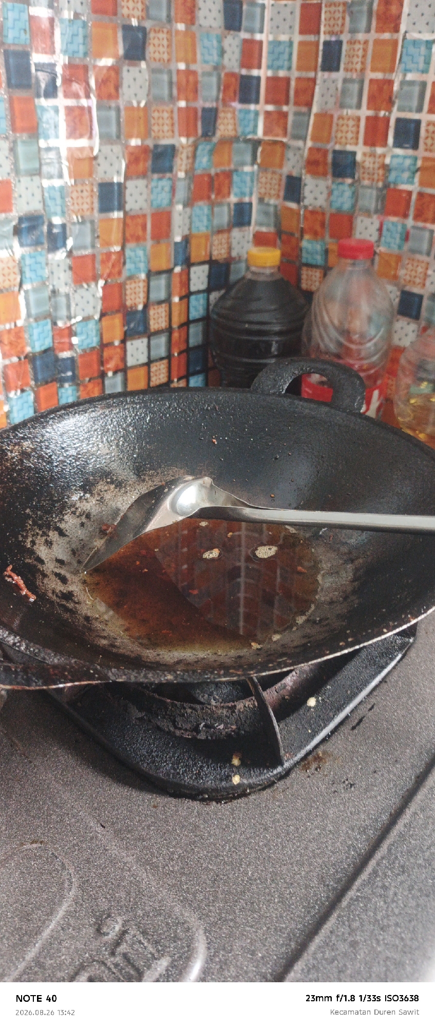 A close-up, slightly overhead shot captures a black cast iron wok filled with oil and a metal ladle resting inside, positioned on a gas stove. The background features a colorful tiled backsplash in shades of orange, blue, and white. To the right of the wok, two bottles are visible; one is a dark bottle with a yellow cap, and the other is a clear bottle with a red cap. The bottom left of the image displays text indicating "NOTE 40 2026.08.26 13:42" and "23mm f/1.8 1/33s ISO3638 Kecamatan Duren Sawit," suggesting the photo was taken in Kecamatan Duren Sawit, East Jakarta, Indonesia. The overall scene depicts a cooking setup with ingredients being prepared.