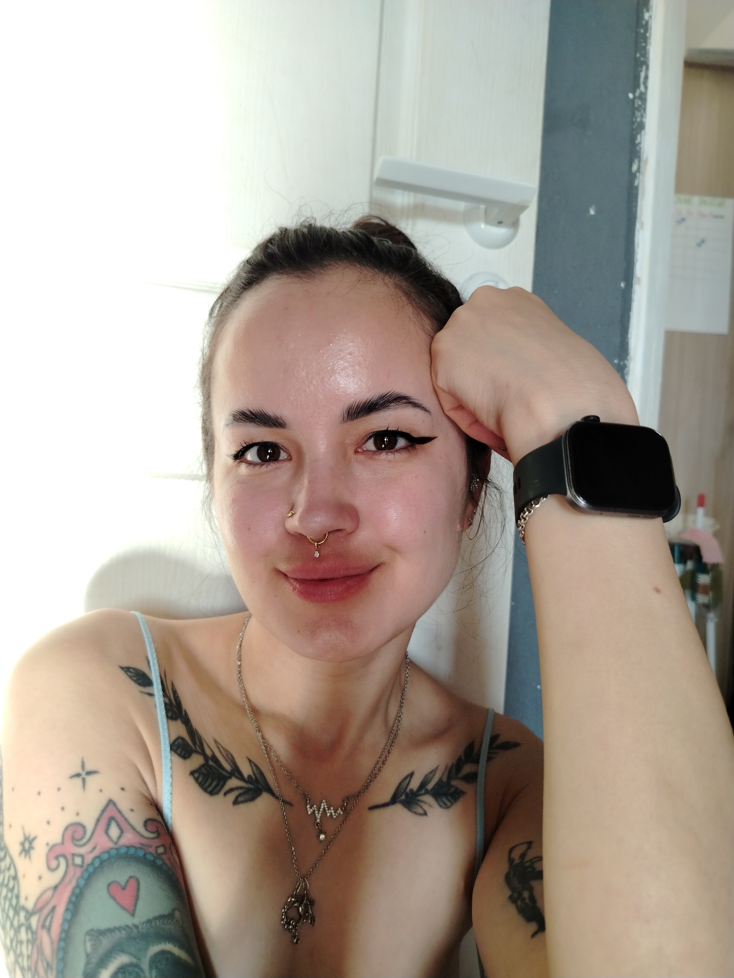 A selfie shows a woman with light skin and dark eyebrows and eyeliner. She has her hair pulled back, and she is wearing a gold nose ring. She is wearing a light blue tank top, and two necklaces. She has tattoos on her chest and left arm. On her left wrist, she is wearing a black smartwatch and a silver bracelet. She is leaning her head on her left hand. The background is a white door, and a sliver of grey wall. There is a small white calendar on the wall behind her. The location is not discernible as being in the city of Noviy, Russia.