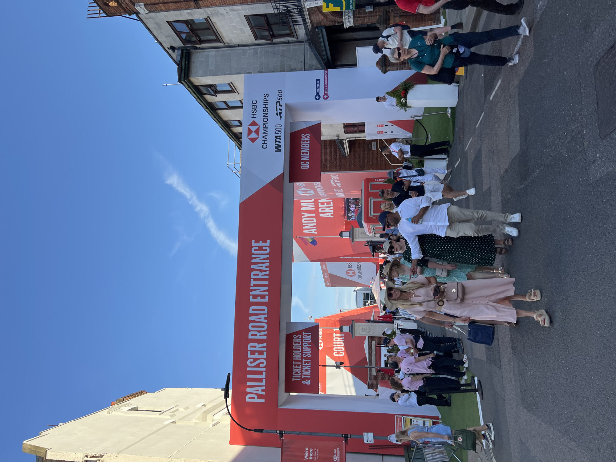 The image shows the "Palliser Road Entrance" to an event, likely a sports tournament, as indicated by the "HSBC CHAMPIONSHIPS WTA 500 ATP 500" signage. A large red and white archway frames the entrance, with the words "PALLISER ROAD ENTRANCE" prominently displayed. Below the arch, a sign reads "TICKET HOLDERS & TICKET SUPPORT." Further signage directs attendees to "QC MEMBERS," "THE VIEW," and "THE CLUBHOUSE."
A group of people, dressed in casual attire and some in event-specific pink shirts, are walking through the entrance. Among them are individuals wearing hats and sunglasses, suggesting a sunny day. Some are carrying bags and purses. The environment appears to be an urban street, with buildings visible on either side of the entrance and a clear blue sky above. A lamppost stands to the left of the archway, and a red barrier is set up on the right. The ground is paved, with white lines indicating a roadway.