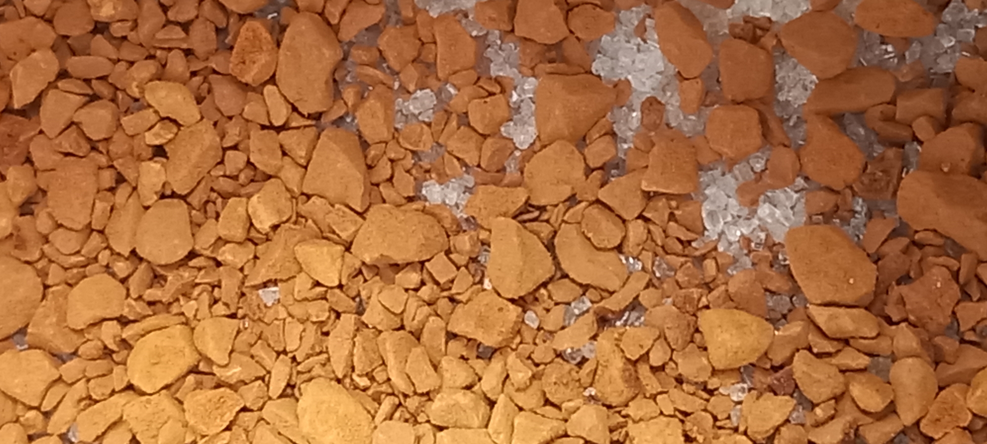 This close-up image displays a granular mixture of two distinct materials. The predominant material consists of irregularly shaped, reddish-brown fragments, varying in size from small crumbs to larger pebbles. These pieces have a dull, earthy, and somewhat porous appearance, resembling crushed iron ore, rust, or a similar mineral.
Interspersed among these reddish-brown particles, particularly visible in the upper and right portions of the frame, are numerous clear to translucent white fragments. These white pieces are generally smaller, with sharp, angular edges, consistent with crushed glass, salt crystals, or other crystalline mineral deposits. The two materials are intermingled, with the smaller white particles often filling the gaps between the larger reddish-brown chunks.
The scene is presented from a top-down perspective, offering a detailed view of the materials' texture and distribution. The lighting is even and diffused, without harsh shadows, highlighting the granular nature of the substances. There is no discernible activity or event taking place, and no people or specific settings are visible beyond the materials themselves. No text is visible in the image.
Given the location context of Kryvyi Rih, Ukraine, which is a major industrial center known for iron ore mining, the reddish-brown material strongly aligns with the appearance of iron ore or its processed forms. The clear/white fragments could be an industrial byproduct, crushed glass from recycling, or another mineral found in the region.