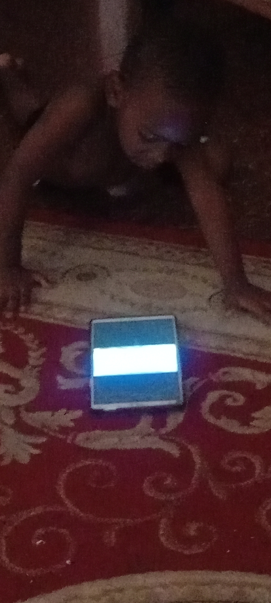 A child is lying on a red rug with a scrolling pattern. The child's face is partially visible and is looking down at a tablet device. The tablet screen is illuminated and shows a horizontal white bar. The child's arms and legs are also visible. The image appears to have been taken indoors in low light conditions. The location in Kano, Nigeria, is not specified in the image or provided context.