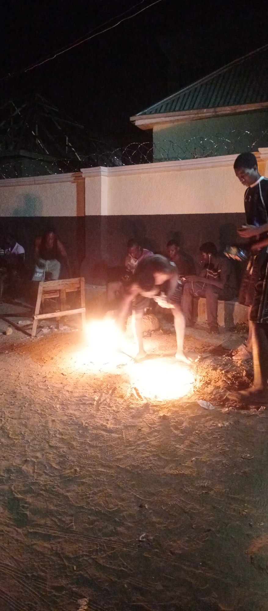 The image captures an outdoor night scene in Keffi, Nigeria, featuring a group of young men gathered around a bright bonfire. The fire, centrally located on sandy ground, emits a strong orange and yellow glow, illuminating the immediate surroundings and casting shadows on the figures.
In the foreground, a barefoot man is bent over, actively tending to the fire, suggesting he is managing the flames or adding fuel. To his right, another young man stands, casually holding what appears to be a mobile phone. In the mid-ground and background, several other men are visible; some are seated on a low wall or bench, and a few also appear to be engaged with their phones or observing the fire. Their faces are partially obscured by shadows or the intensity of the firelight.
A rustic wooden stool or small table is positioned near the fire on the left side. Behind the group, a light-colored wall topped with coils of barbed wire marks a boundary, and above it, part of a building with a dark green, possibly corrugated metal, roof is visible against the dark night sky. The time of day is clearly night, with the bonfire acting as the primary light source. The setting implies a casual social gathering or communal event. No visible text is present in the image.