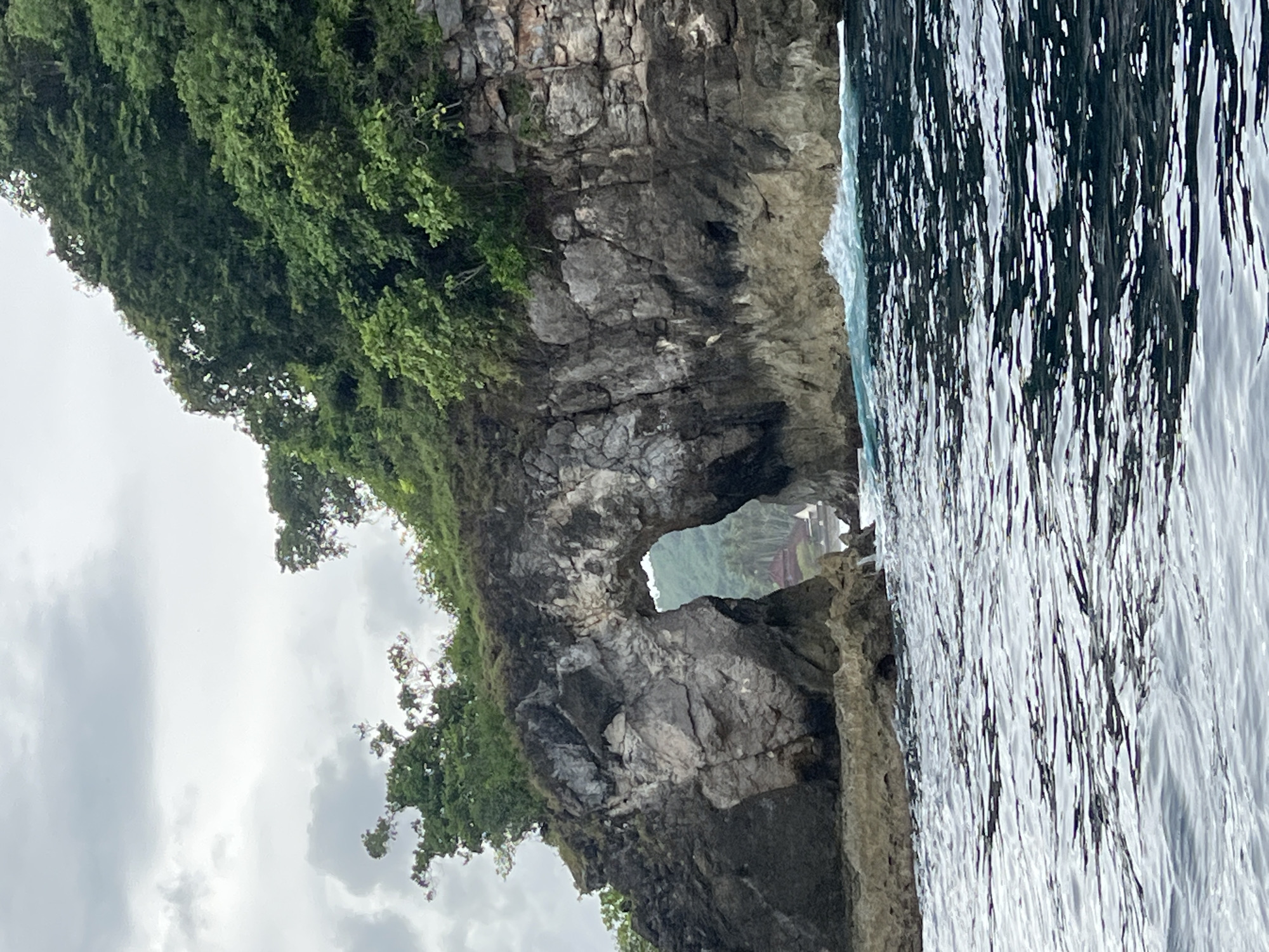 An image captures a natural rock archway carved into a rugged cliff face, set against a body of water and a cloudy sky. The primary subject is a large, dark grey rock formation, partially covered by dense, lush green vegetation on its upper slopes. A prominent natural arch, or sea cave, pierces through the lower part of the cliff, framing a distant view.
Through the archway, glimpses of what appears to be another landmass or island are visible, also covered in greenery, with faint suggestions of reddish-brown structures (possibly rooftops) nestled among the trees. To the right of the cliff, dark, reflective water extends, showing rippled textures and highlights from the sky. On the left side of the image, an overcast sky filled with soft grey clouds dominates the upper background.
The scene depicts a serene coastal landscape, likely viewed from a boat. The light suggests an overcast day, possibly late morning or afternoon. The location is Nusa Penida, Indonesia, renowned for its dramatic natural beauty and rock formations. There are no people or explicit activities taking place, and no visible text. The overall impression is one of natural grandeur and tranquility.