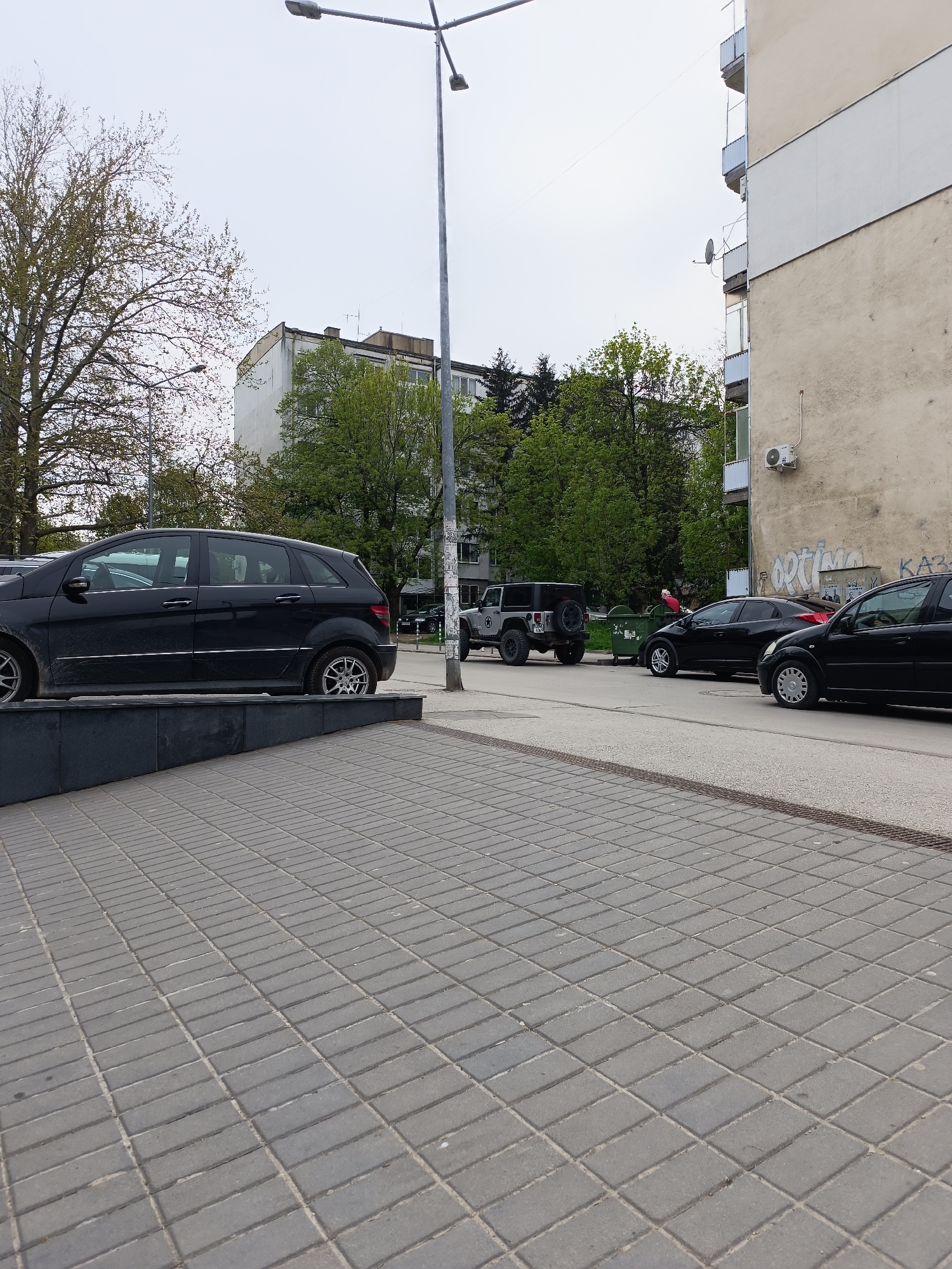 This media file captures an urban street scene in Pleven, Bulgaria, under an overcast daytime sky. The foreground is dominated by a gray paved sidewalk, exhibiting a diagonal brick pattern towards the right. A low, dark gray concrete barrier separates the sidewalk from the street on the left.
The street itself features several parked vehicles. On the left, a black Mercedes-Benz B-Class hatchback is parked, partially obscured by the barrier. Further down the street, a black Jeep Wrangler with a visible rear-mounted spare tire is positioned near a large green dumpster. A person wearing a red top is faintly visible leaning into or near the dumpster. Beyond the dumpster, two more black cars are parked: a Honda Civic hatchback (distinguished by its red taillights) and a smaller black hatchback, possibly a Citroën C3.
In the background, large trees with light green, early spring foliage line the street. Behind the trees, several multi-story apartment buildings are visible. The building on the right features balconies with metal railings, a satellite dish, and an external air conditioning unit. Its facade shows signs of age and includes visible graffiti, with "OPTIMA" and Cyrillic characters like "KA3" being discernible. A prominent gray lamppost stands near the center of the image, extending its arms across the upper portion of the frame. The overall scene depicts a typical residential street.
