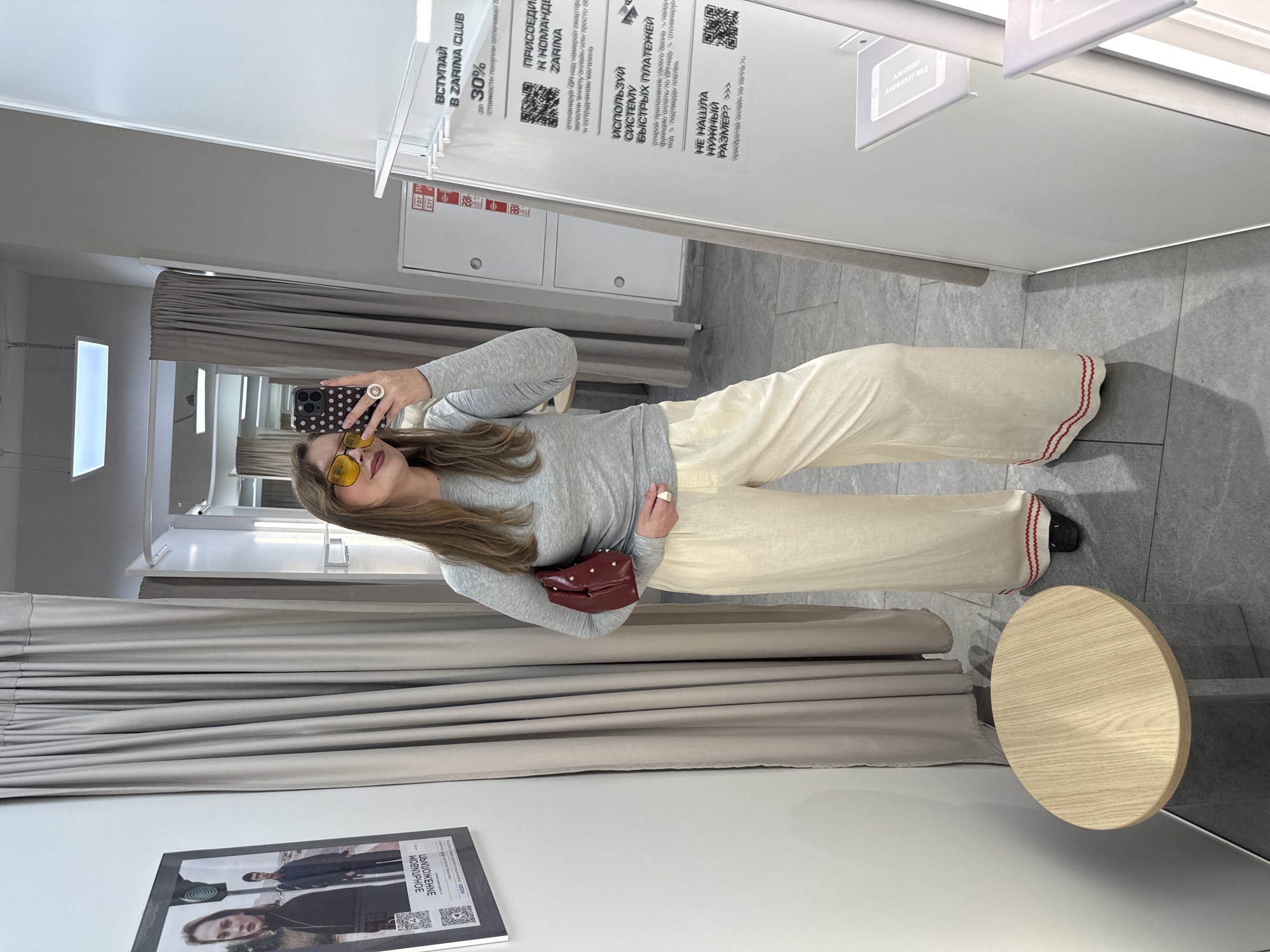 Here's a matter-of-fact description of the image:
**Scene:** A dressing room, potentially in a retail store. It appears to be taken in Izhevsk, Russia, based on the Cyrillic text on signage.
**People:** One woman is visible, holding a phone and taking a selfie in a mirror. She's wearing yellow-tinted sunglasses, a long-sleeved grey top, and light-colored pants with a red and white accent at the hem. She's holding a small, dark red, studded handbag.
**Objects:**
* A mirror reflects the woman and the dressing room interior.
* A round wooden stool is visible.
* Clothing racks and curtains line the walls of the dressing room.
* Signage with text in Russian is mounted above the racks. The signs advertise a "ZARINA Club" and other promotional information.
* A poster/advertisement with imagery of people is on the wall.
* The woman's smartphone features a polka dot design.
**Actions:** The woman is taking a picture of herself in the mirror.
**Environment:** The dressing room has grey-toned walls and tiled flooring. The lighting seems bright. The atmosphere suggests a retail environment.