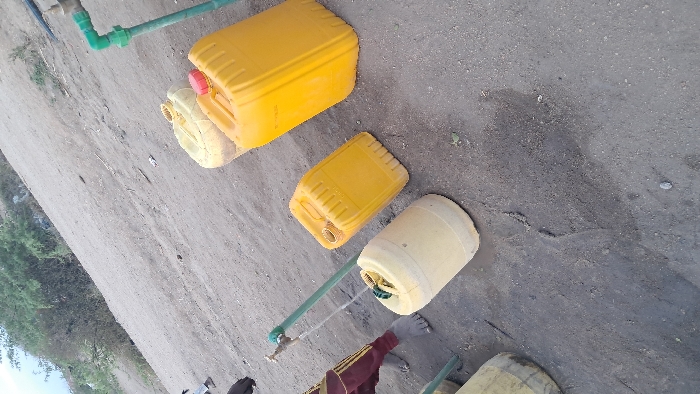 The scene features several yellow and beige plastic containers, likely jerrycans, arranged on a gray surface. A green pipe with a spigot is visible, seemingly used for filling the containers with water. One can is capped with a red lid. A person wearing a maroon and gold sleeve is partially visible near the bottom of the frame, suggesting someone is attending to the water-filling process. The backdrop reveals a dusty path and sparse greenery.