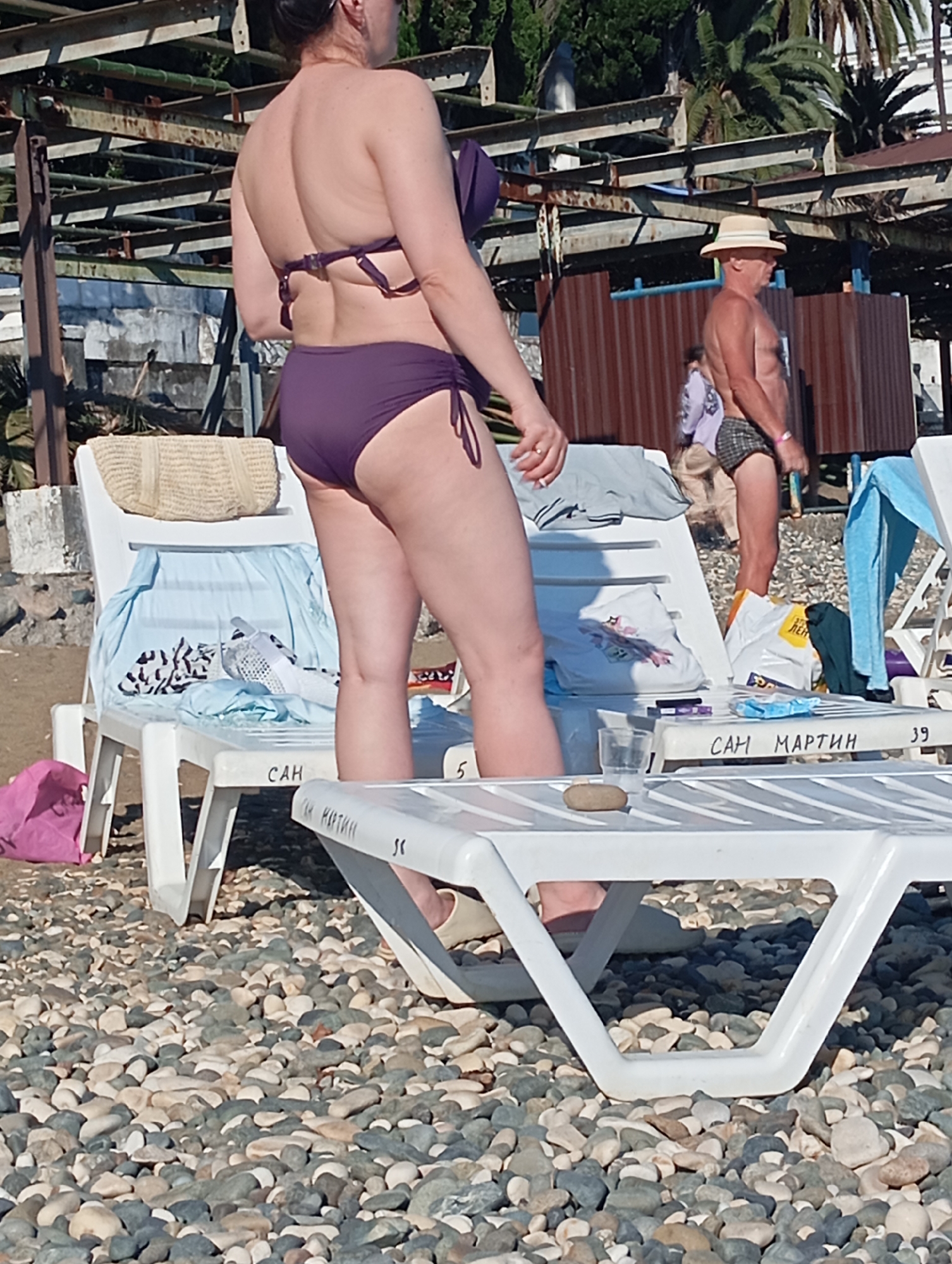 The image depicts a beach scene in Sokhumi, Abkhazia, characterized by a pebble-covered ground and rows of white plastic lounge chairs. In the foreground, a woman wearing a purple bikini is partially visible, bending over. Next to her, on a lounge chair, is a woven straw bag and a light blue towel draped over it. Another lounge chair to the right has a light blue towel hanging from it and various items including shopping bags and a glass. In the background, a man in checkered swim trunks and a straw hat stands near a wooden structure. Palm trees and other foliage are visible behind the structures, suggesting a warm, outdoor environment. Some of the lounge chairs have lettering and numbers written on them, possibly indicating locations or rentals.