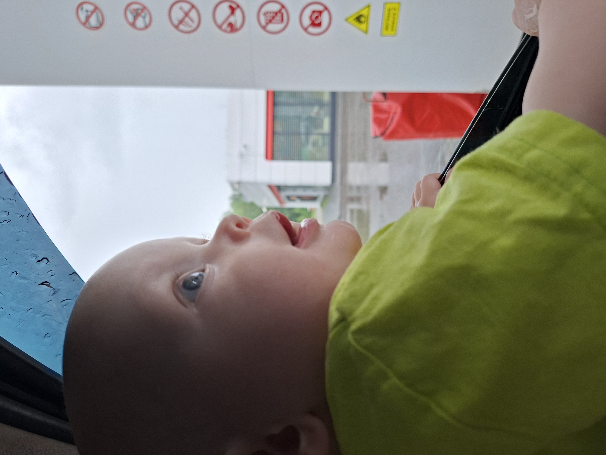 An extreme close-up, low-angle shot shows a bald baby looking out of a car window. The baby is wearing a bright green t-shirt. Rain streaks down the window, and the blurry background shows a gas station with red and white buildings. There are several red circular prohibition signs on a white post to the right of the baby. The scene is likely taking place at a gas station in Krilovo, Russia, during a rainy day.