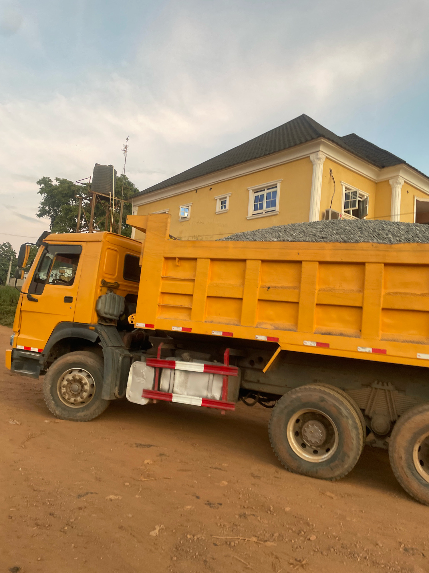 In the city of Abuja, Nigeria, a bright yellow dump truck is parked on a dirt road. The truck is loaded with a substantial amount of grey gravel. The truck's bed is clearly visible, showing its construction and the material it carries. To the right of the truck, a two-story yellow building with a dark grey roof stands, featuring several windows, some with blue panes. The sky above is partly cloudy with a light blue hue. Behind the truck and to the left, a large green tree is visible, and a water tank supported by a metal structure can be seen. The scene appears to be a construction or material delivery site.