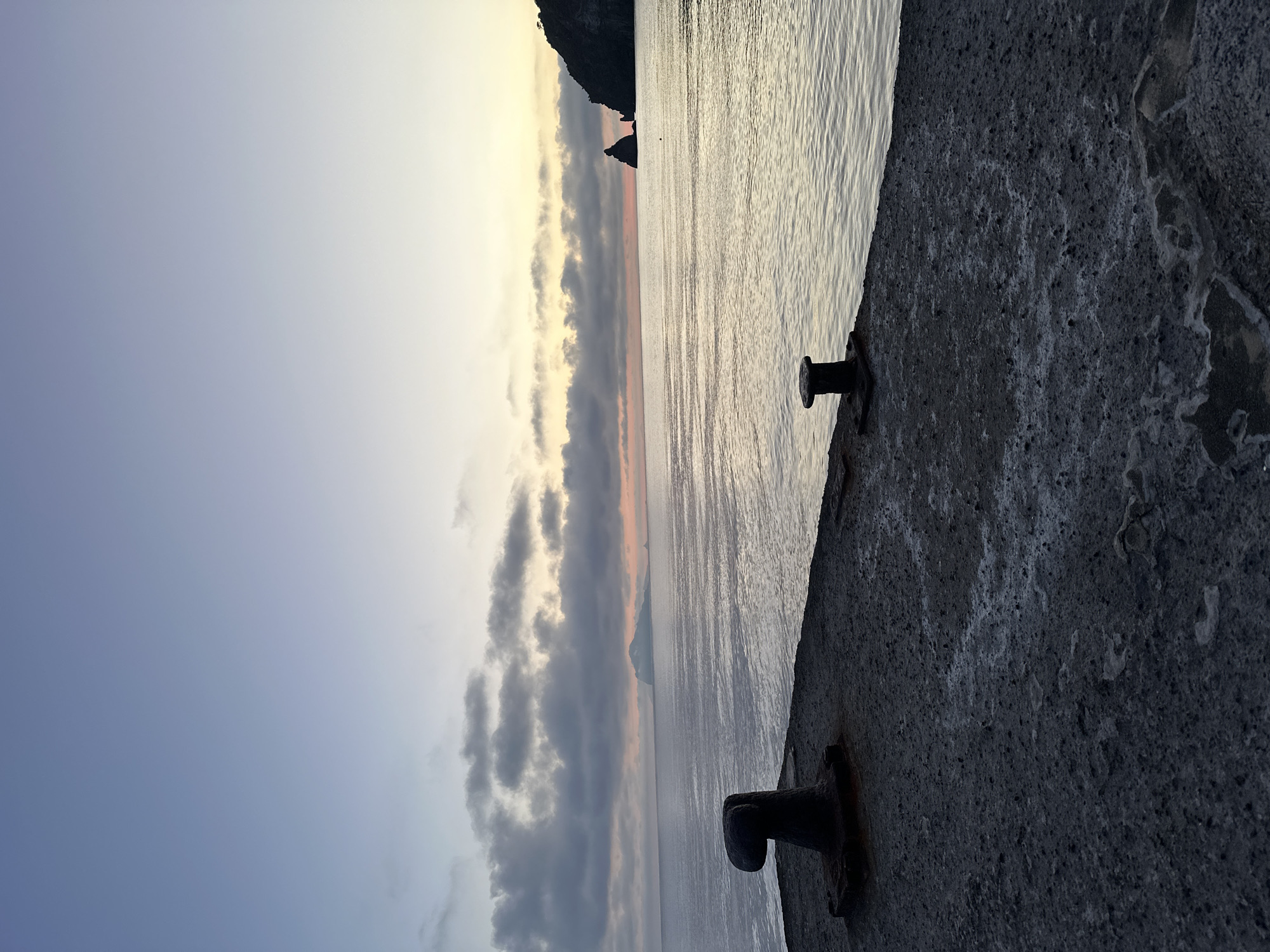This media file captures a tranquil coastal scene in Lipari, Italy, likely during either sunrise or sunset. The image is composed with a strong horizontal division: the textured, dark grey concrete surface of a pier or dock occupies the right foreground and mid-ground, extending vertically in the original orientation. Two weathered metal mooring bollards are prominently featured on the concrete, appearing rusted in places.
To the left of the concrete, the calm, silvery-grey water of the sea stretches towards the horizon, reflecting the soft, warm light from the sky. Gentle ripples are visible on the water's surface, indicating a lack of strong wind.
The sky above displays a gradient of colors, transitioning from a soft blue-grey at the top to a pale yellow and orange near the horizon, where the sun's presence is implied just beyond the frame. Several long, dark, horizontal clouds are spread across the mid-sky, providing contrast against the softer hues. In the far distance, the dark silhouette of a mountain or landmass is faintly visible against the brighter horizon.
No people or explicit activities are depicted, contributing to a sense of peacefulness and serenity. The overall impression is one of quiet contemplation at the beginning or end of the day. No visible text is present in the image.