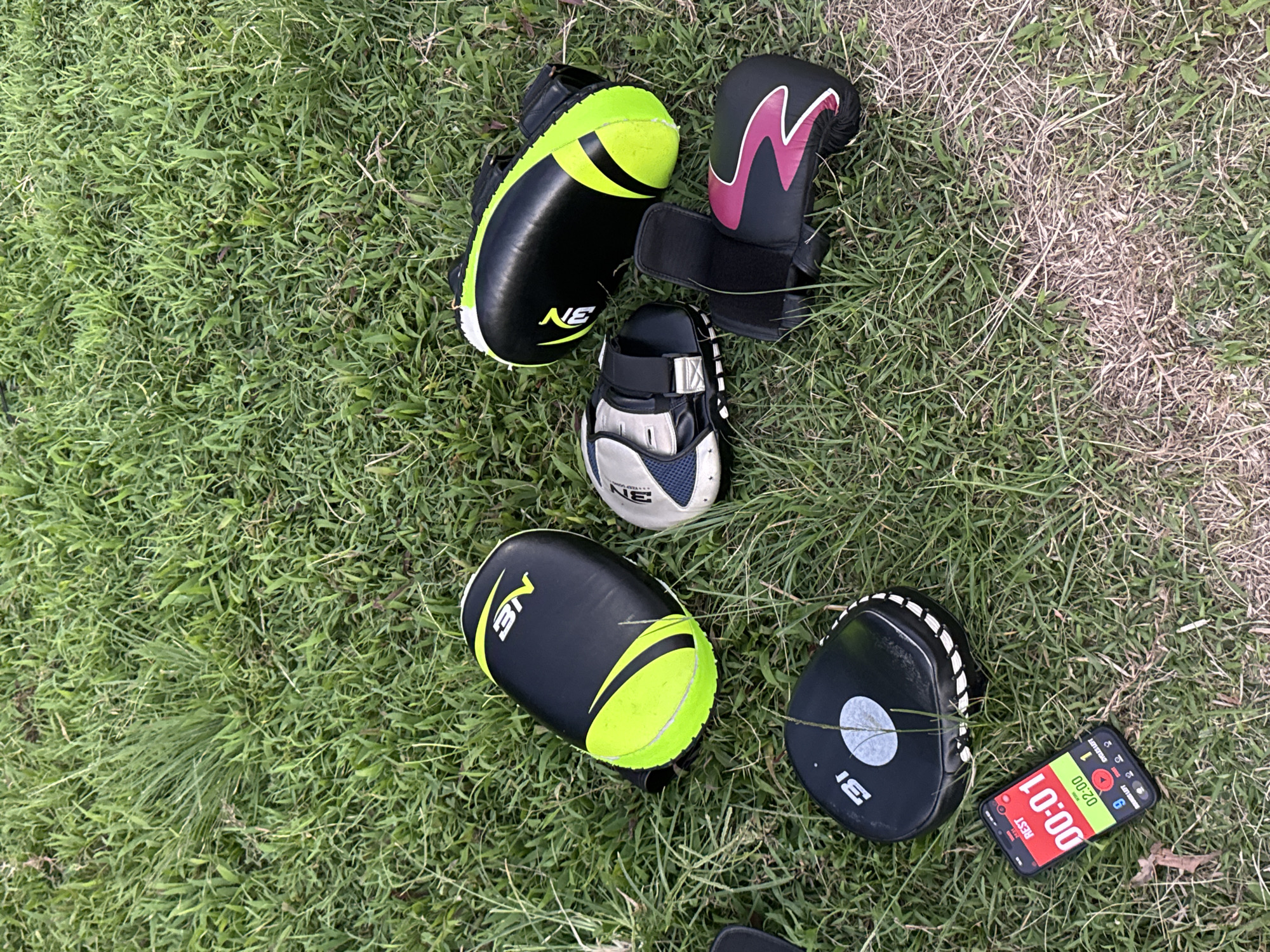 The image shows several items of boxing training equipment laid out on a patch of grass. There are two black and neon green punch mitts, one white and blue hand protector, and a black hand mitt with a pink and black design. A smartphone displaying a timer is also on the grass. The background is a dense covering of green grass with some dry patches. There are no people visible in the image. The scene appears to be an outdoor training area.