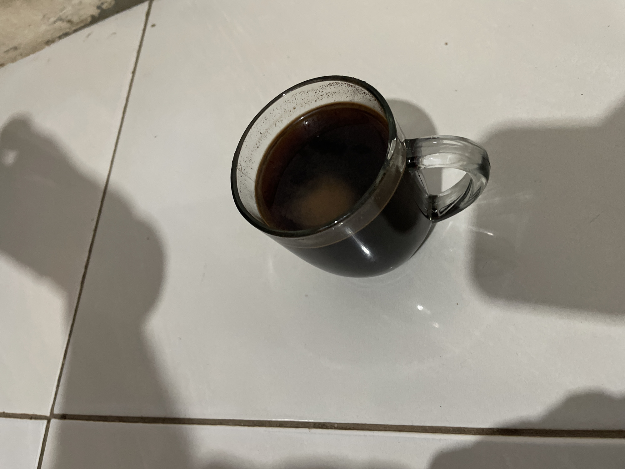 A glass mug filled with dark liquid, possibly coffee, is tilted on a white tiled floor. The mug has a clear glass handle and is casting a shadow. The floor appears to be sectioned by grout lines. The lighting suggests an indoor environment, possibly a home or cafe, in Kertosari, Indonesia. The overall scene is still and uneventful.