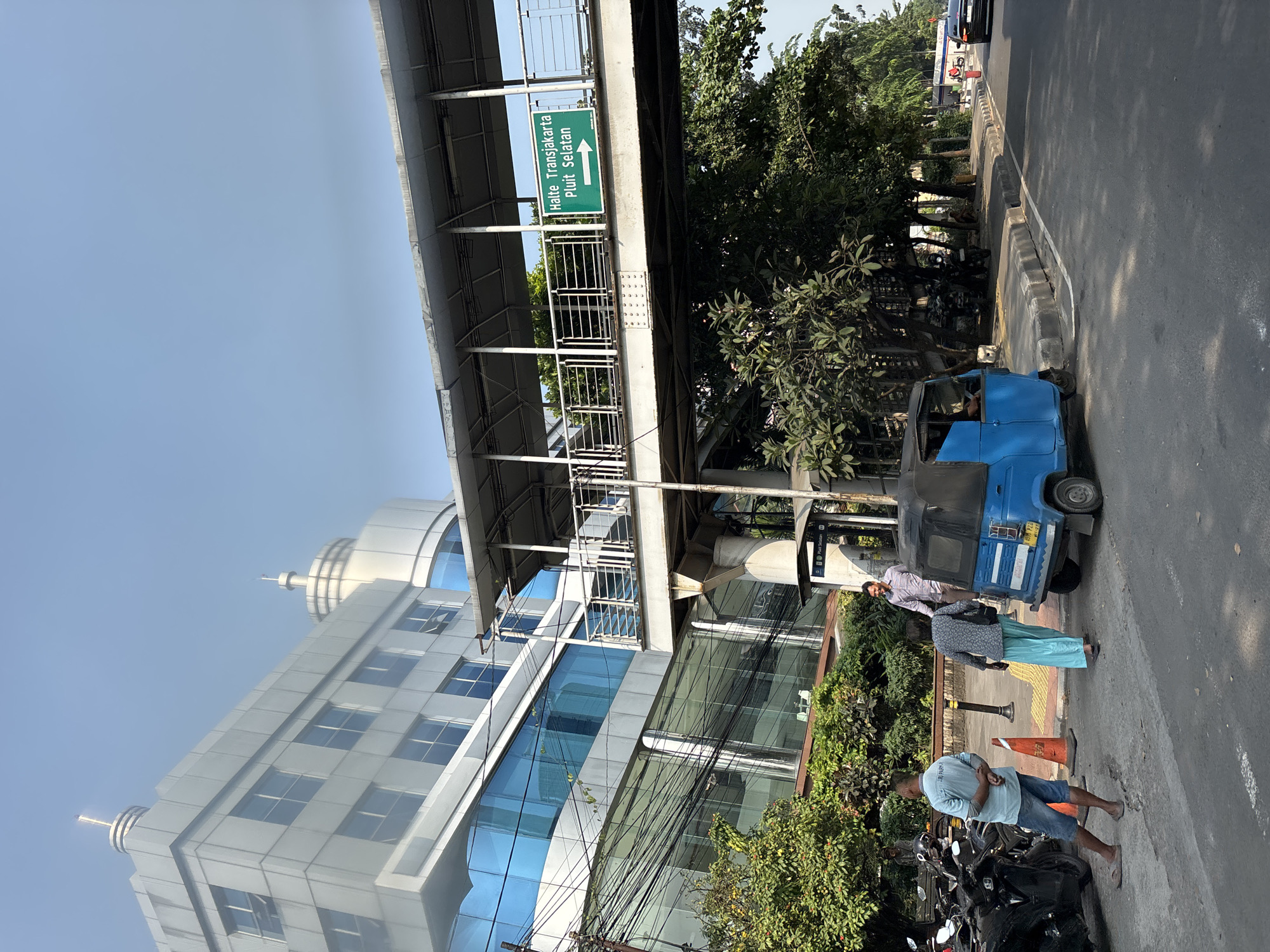 The image depicts a street scene in North Jakarta, Indonesia, specifically near Halte Transjakarta Pluit Selatan. In the foreground, a blue auto-rickshaw (bajaj) is parked on the side of a road, partially obscuring a group of parked motorcycles. Three people are visible: a man in a light blue shirt and denim shorts stands with his back to the camera, looking towards the parked motorcycles. A woman in a patterned top and a long blue skirt is walking away from the auto-rickshaw. A man in a patterned collared shirt and dark pants is standing further down the sidewalk, facing the woman.
An elevated pedestrian walkway, part of the Transjakarta bus rapid transit system, spans the road. A green sign on the walkway reads "Halte Transjakarta Pluit Selatan" with a directional arrow pointing to the right. Behind the walkway, a modern, multi-story building with blue glass windows and metallic exterior is prominent. Lush green trees and foliage line the sidewalk and the base of the building. Overhead, a network of electrical wires is visible against a clear blue sky. The road surface is asphalt, with a distinct curb separating it from the sidewalk. The overall impression is of a bustling urban environment during daylight.