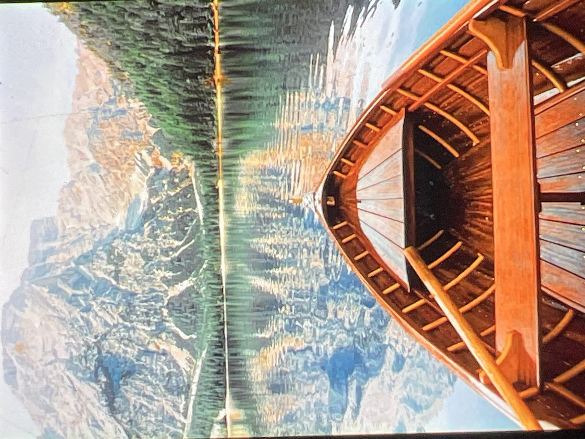 The image shows a wooden rowboat with an orange hue, facing forward on a calm body of water. The boat's interior is visible, with wooden slats forming the floor and curved ribs along the sides. A single oar rests across the boat. The water reflects the surrounding landscape, which consists of a steep, rocky mountain range with patches of snow and a dense forest of green pine trees bordering the water's edge. The sky is bright, with hints of white and a light glow. The overall scene depicts a serene natural landscape viewed from the perspective of someone in a boat. The location is identified as Unknown, Unknown.