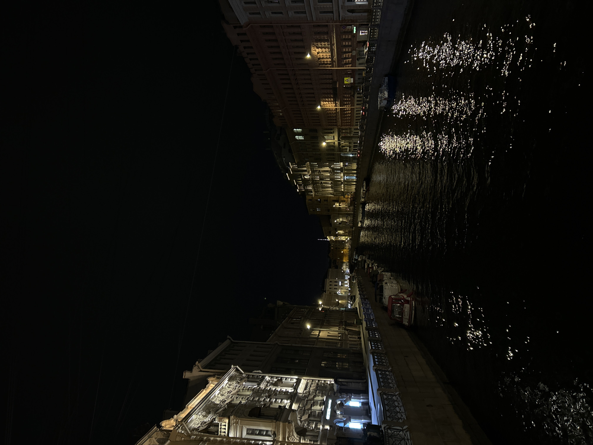 At night, a canal in Saint Petersburg, Russia is framed by illuminated buildings. Streetlights reflect on the water. Cars park along the canal.