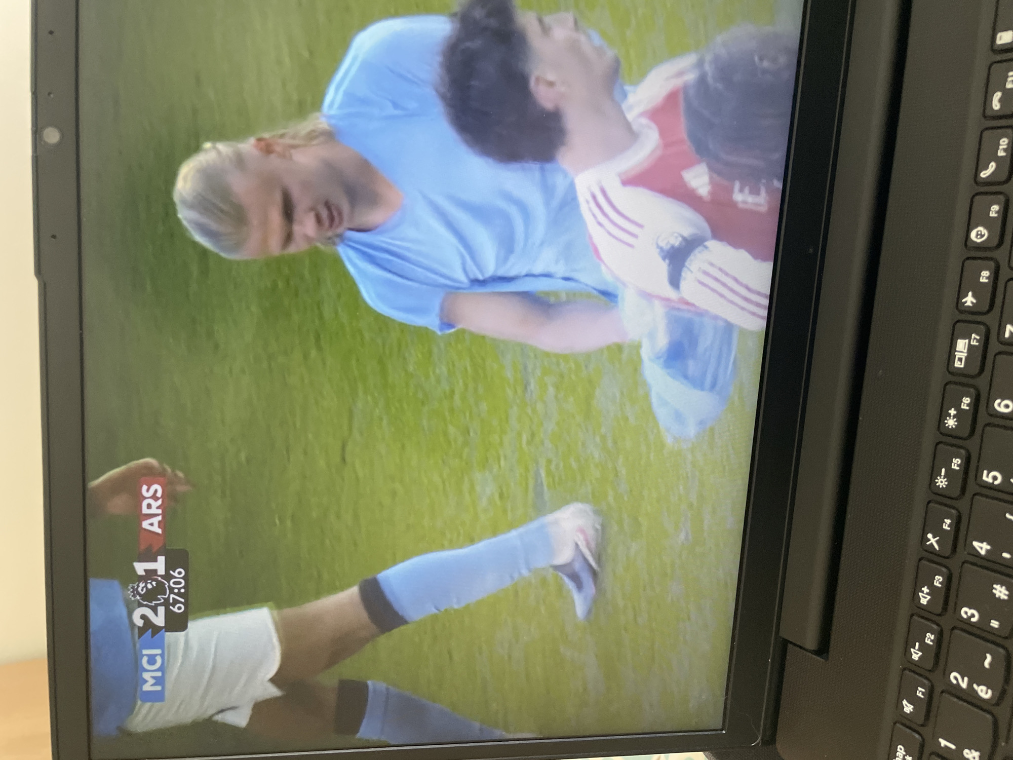 The image shows a laptop screen displaying a football (soccer) match between Manchester City (MCI) and Arsenal (ARS). The score is MCI 2-1 ARS, with the game clock at 67:06. On the screen, two players are depicted on the field: one in a light blue Manchester City jersey, who appears to be Erling Haaland, and another in a red and white Arsenal jersey. The setting is a grassy field, presumably a football pitch. The photo is taken in an indoor setting (as suggested by the presence of the laptop). The location in the city of Parthenay, France, is not indicated or verifiable from the content of the image. The keyboard of the laptop is visible.