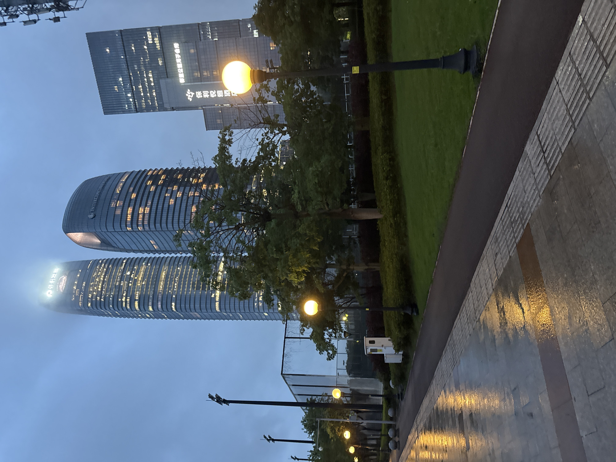 The image depicts a modern cityscape at dusk, likely in Haizhu District, China, characterized by towering glass skyscrapers illuminated from within. In the foreground, a wet, paved walkway reflects the warm glow of streetlights. A dark asphalt path runs alongside the walkway, bordered by a neatly manicured green lawn. Several lampposts, their globes emitting a soft yellow light, punctuate the path. Behind the lawn, a line of trees with lush green foliage stands against the backdrop of the buildings. One of the skyscrapers prominently features the "InterContinental" logo. Another building displays Chinese characters which translate to "Poly Kosa Club" and "Poly Kosa Hotel." The sky is overcast, creating a muted, twilight atmosphere. The scene suggests an urban park or public space adjacent to commercial buildings, possibly after a rainfall given the wet pavement. No people are visible in the frame.