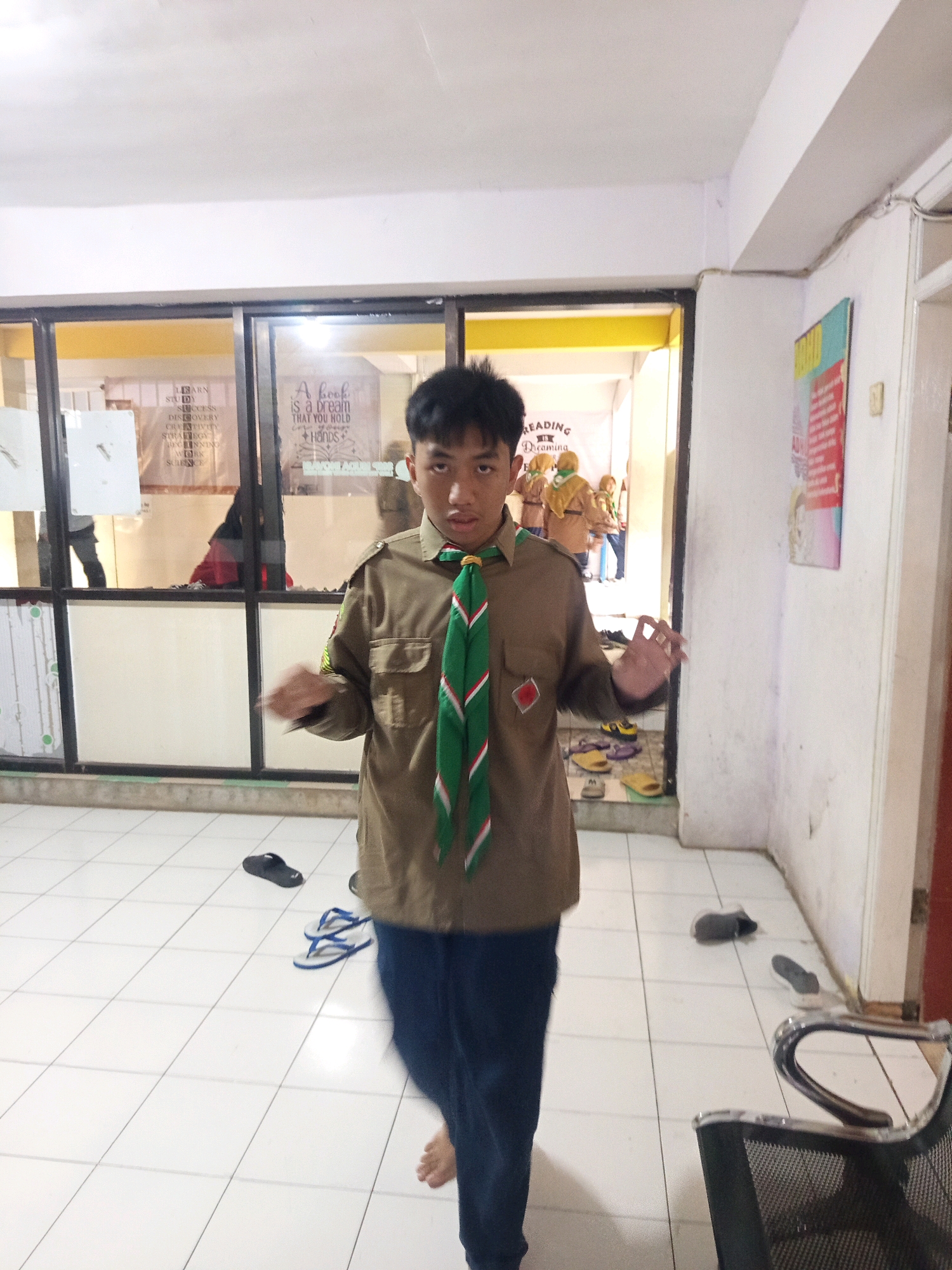 A young Indonesian male, dressed in a scout uniform consisting of a brown long-sleeved shirt and dark blue trousers, stands indoors on a white tiled floor. He wears a green neckerchief with red and white stripes. The background features large glass doors revealing an interior space with yellow walls. Posters with text are visible on the walls inside. To the right of the main subject, a white wall with a red and white poster is visible, and a metal chair with a mesh seat is partially in frame. Several pairs of flip-flops are scattered on the floor around the subject. The lighting appears to be artificial. The scene is likely within a school or community building in Malang, Indonesia.