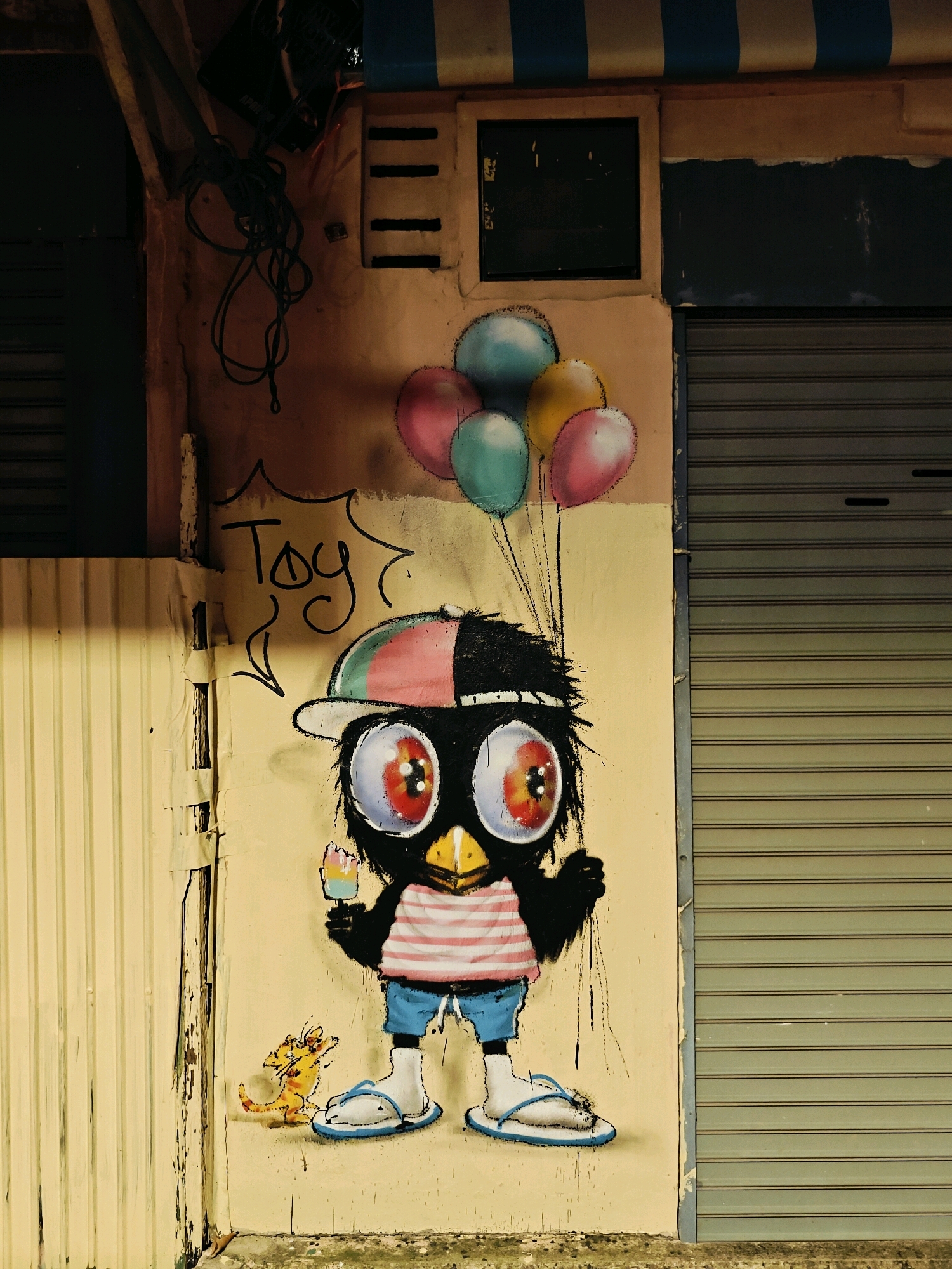 The image captures a colorful graffiti mural on a textured wall, likely in an urban setting. The mural depicts a cartoonish black bird character with large, wide eyes, wearing a cap, a striped shirt, and shorts. The bird is holding a popsicle and balloons, and the word "Toy" is scrawled above it in black ink, with an arrow pointing down towards the character. To the left of the bird, a small yellow cat-like creature is painted. The wall itself shows signs of age and weathering, with peeling paint and visible textures. In the background, a metal shutter, typical of shops or buildings in Bangkok, Thailand, is visible on the right, and a section of corrugated metal sheeting is on the left. The overall lighting suggests it may be evening or a dimly lit alley. The scene appears to be a snapshot of street art in a bustling city environment.
