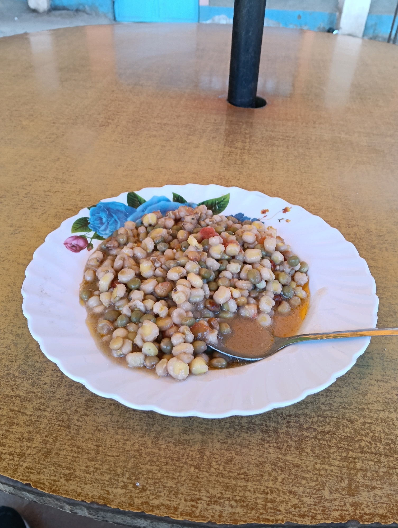 A plate of cooked peas and corn is positioned on a wooden table. The plate has a decorative floral pattern featuring blue and pink flowers. A silver spoon rests in the food, partially submerged. The background shows a blue wall and a black pole. The scene is likely located in Zombe/Mwitika ward, Kenya, as indicated by the user's prompt, suggesting an outdoor or semi-outdoor eating area.