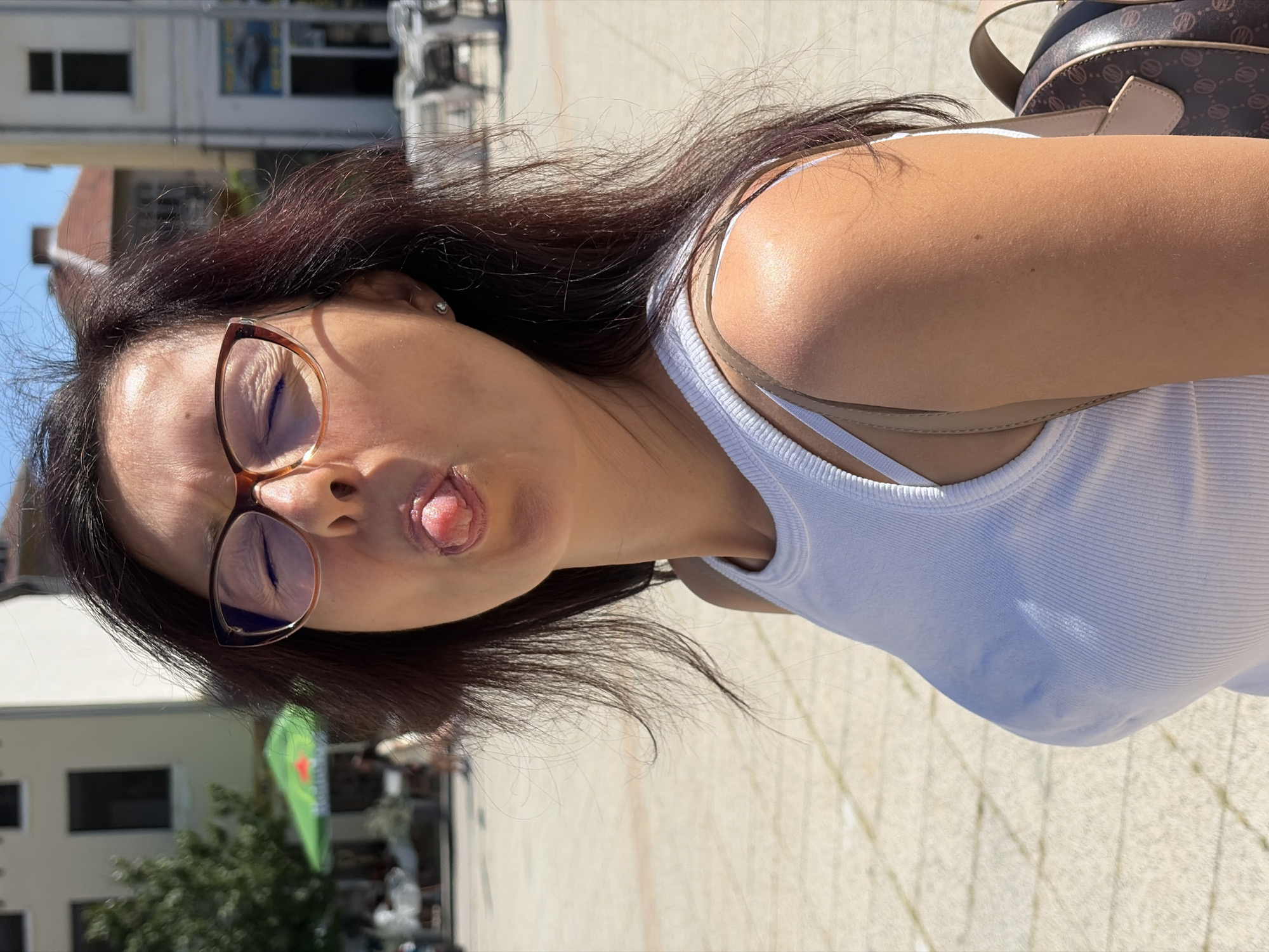 A medium shot photograph captures a woman sticking her tongue out at the camera, her eyes closed. She is wearing a white tank top and brown-rimmed glasses. The background suggests an outdoor urban setting with buildings and paved ground. A green umbrella and people are visible in the distance, indicating a public square or street in Vratsa, Bulgaria.