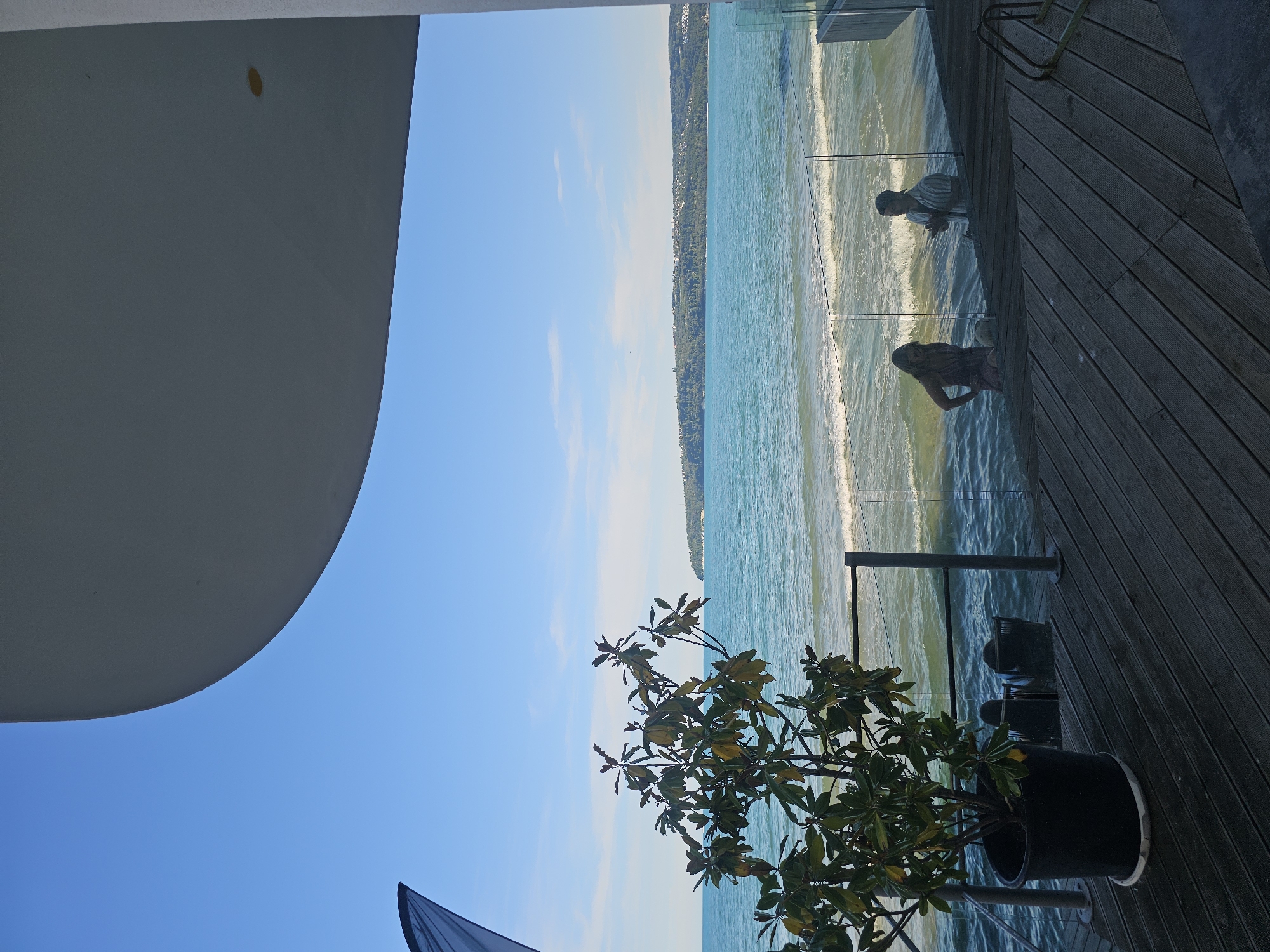 A sunny day with clear blue skies is depicted from a balcony overlooking a tranquil ocean. The scene is bathed in bright sunlight, suggesting it's daytime. A large, curved white structure dominates the left side of the frame, possibly a balcony roof or part of a building's architecture. Below it, a potted plant with green leaves adds a touch of nature to the foreground.
Beyond the balcony, a vast expanse of turquoise water stretches out, meeting a distant shoreline lined with trees and buildings. Gentle waves roll onto the shore, creating a soothing visual. A wooden deck, with planks running diagonally, forms the right side of the foreground, leading to a glass railing.
Reflected in the glass railing are two figures. One, in the upper reflection, appears to be a person in a striped shirt, possibly looking out at the sea. Below them, another figure, silhouetted against the bright water, is also visible. Their presence suggests a peaceful, relaxing atmosphere. The overall mood is serene and calm, with a hint of vacation or leisure. No visible text is present in the image.