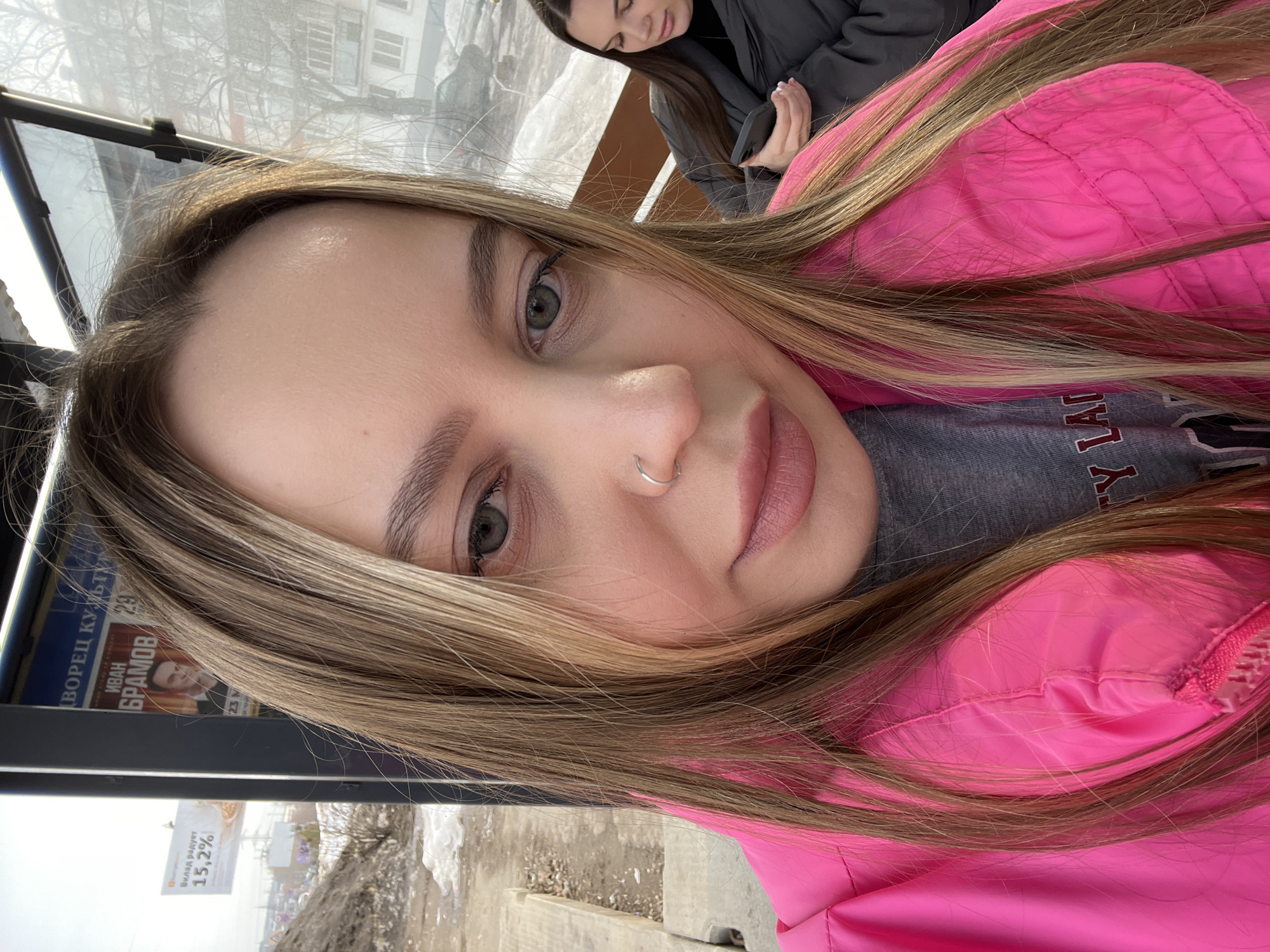 An outdoor scene captures two adult females within a bus stop shelter in Chaikovsky, Russia, during what appears to be a late winter or early spring day, indicated by patches of melting snow and slush on the ground.
In the foreground, a woman with long, light brown and blonde hair, light-colored eyes, and a silver nose ring on her left nostril is shown in a close-up, selfie-style shot. She is wearing a bright pink padded jacket over a grey t-shirt and is looking directly towards the camera.
To the right and slightly behind her, a second woman with dark hair and a dark grey jacket is seated on a bench inside the shelter, looking down at and operating a black smartphone held in both hands.
The bus shelter features clear glass panels and a dark metal frame. Through the glass, two large advertisements are visible:
1. Immediately behind the foreground woman, a poster within the shelter's frame displays Cyrillic text, partially legible as "ДВОРЕЦ КУЛЬТУРЫ" (Palace of Culture) and "ИВАН БРАМОВ" with an accompanying photograph, suggesting a cultural event or political campaign related to a local Palace of Culture.
2. Further in the background, to the left, another large billboard is partially visible, showing "ВКЛАД РАДУГА" and "15,2%".
Beyond the bus stop, the environment includes a large mound of earth or debris to the left, leafless trees, and multi-story residential buildings. A dark car is visible parked on the street. The sky is bright, suggesting a clear or lightly overcast day.