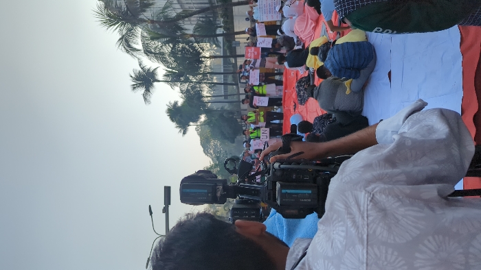 A group of people sit on a red and white cloth while protesting. Some hold signs. They are facing a street lined with palm trees and buildings. In the foreground, a cameraman with a professional video camera is recording the scene. The camera is black with blue accents. The cameraman is wearing a light-colored shirt with a floral pattern. The sky is bright and hazy.