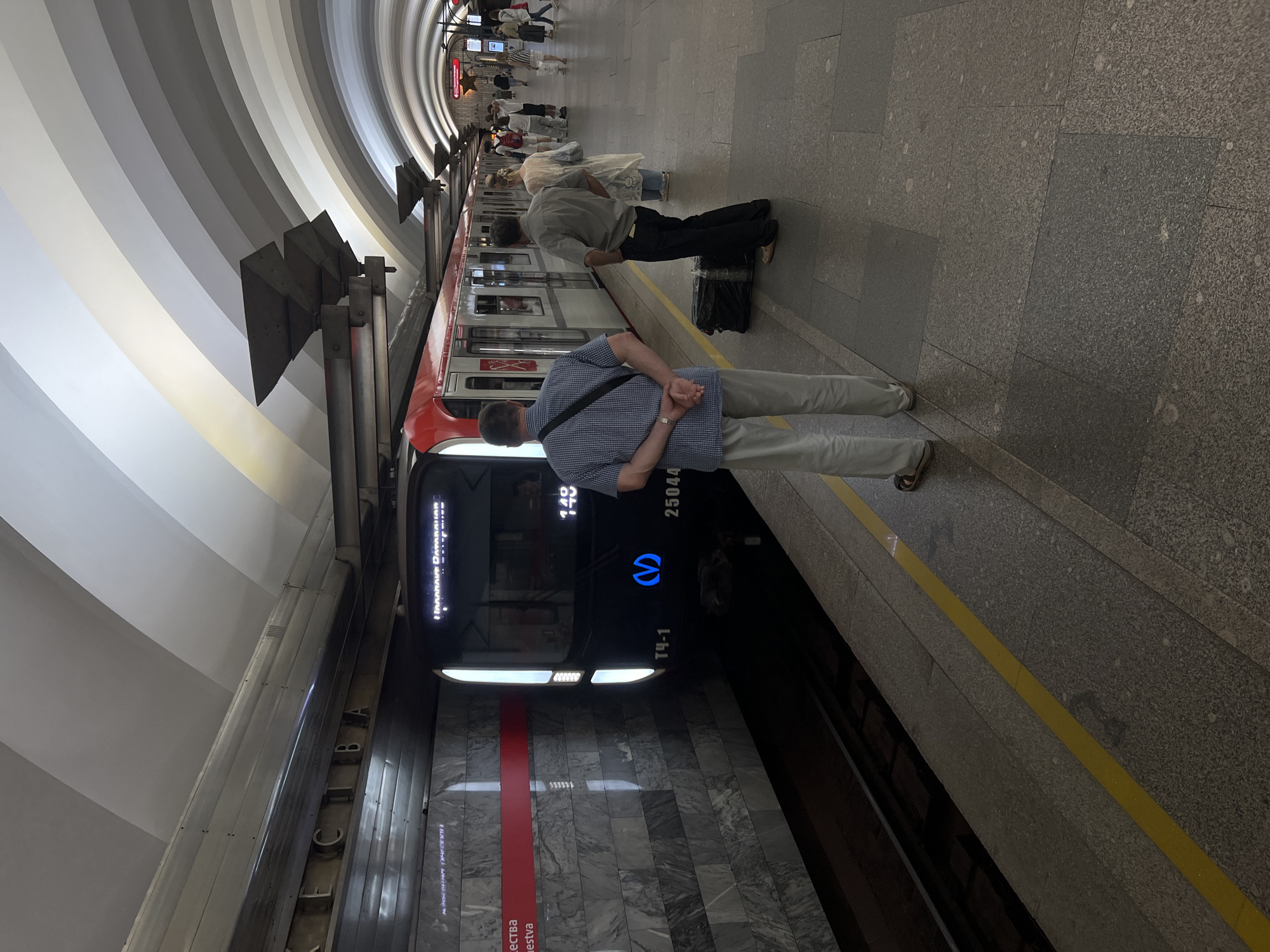 The image depicts a metro station in Saint Petersburg, Russia. A modern red and white train is stopped at the platform, with its doors open. Several passengers are visible, some waiting to board, others standing on the platform. One man in a checkered shirt stands with his back to the camera, his hands clasped behind him. Another man in a grey shirt stands with his hands in his pockets, next to a large black trash bag. The station features a distinctive arched ceiling with alternating bands of white and grey, and the platform is lined with a yellow safety stripe. The station name "ЕЛИЗАВЕТИНА" (Elizavetina) is visible on a sign above the platform. The lighting is somewhat dim, typical of an underground metro environment.
