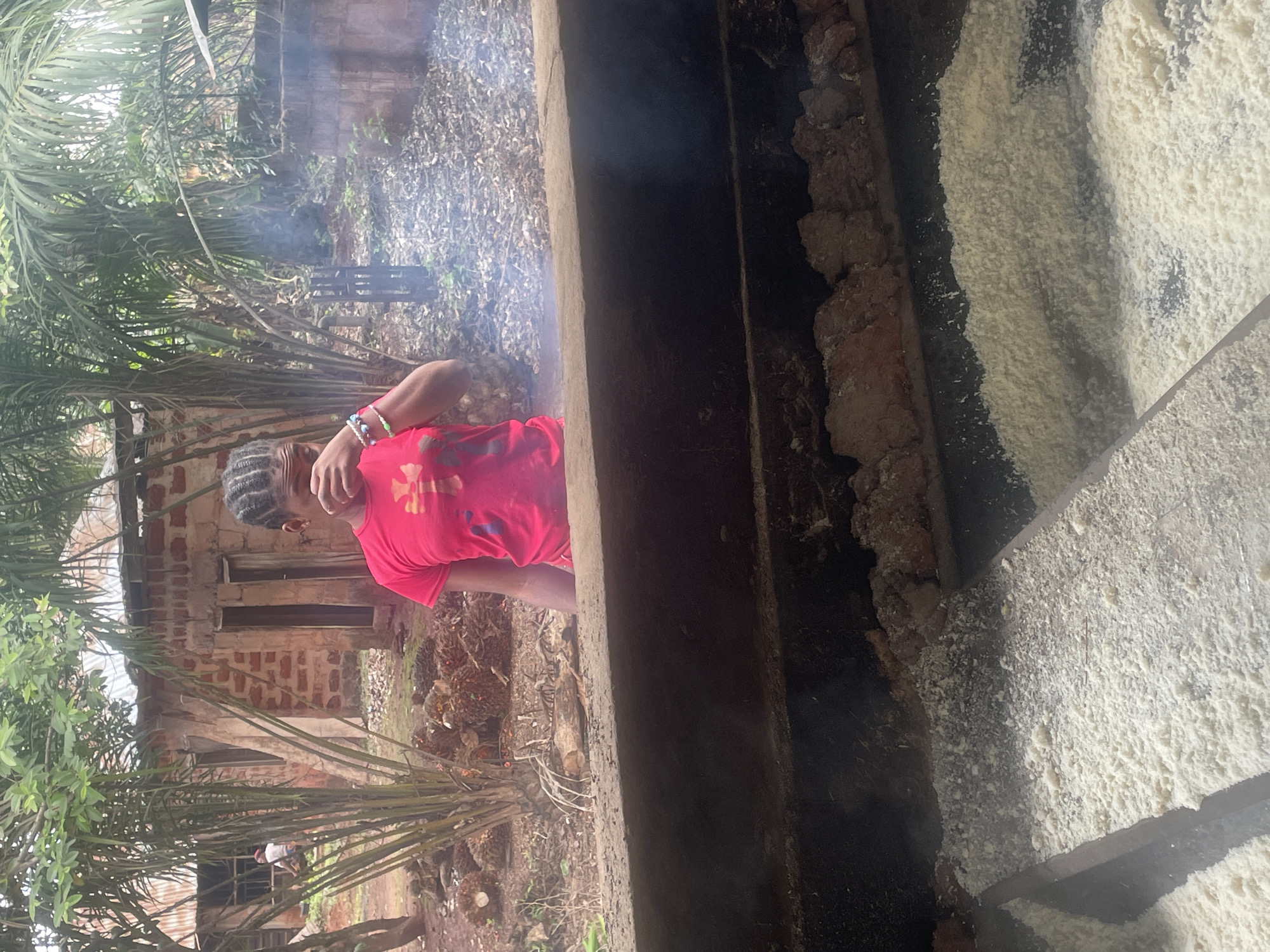 A young woman in a red t-shirt and bracelets stands near what appears to be a large container or structure. The location appears to be outdoors, with a building made of red brick with window, and a variety of vegetation including palm trees and other foliage visible in the background. There is smoke rising in the background. The scene is in Eke, Nigeria. A small white figure is on the corner of the building.