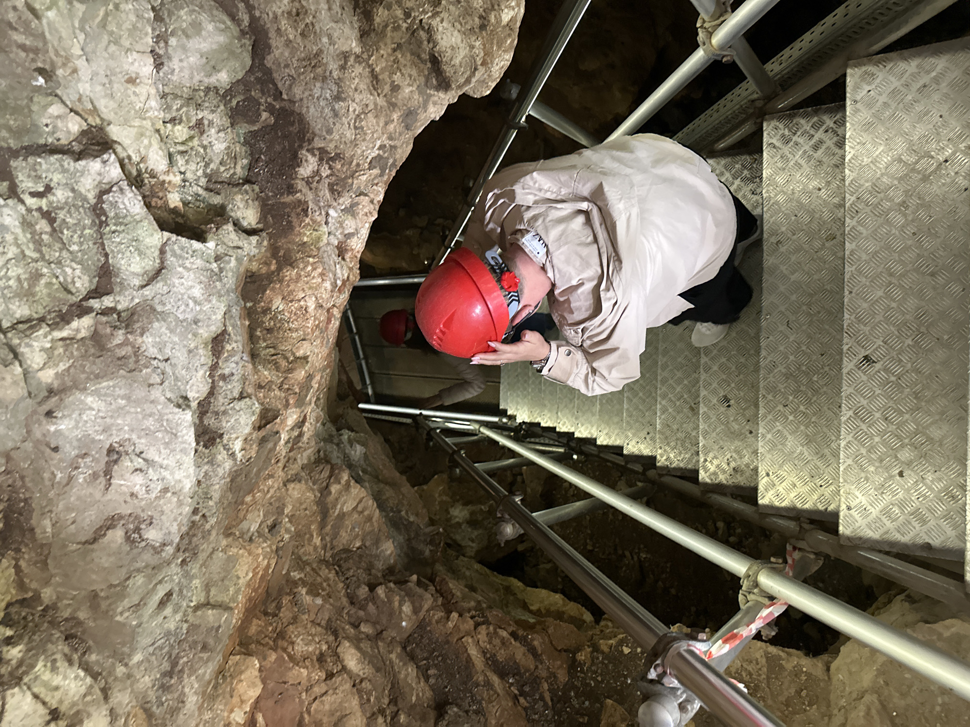 A high-angle shot looking down into a cave with metallic stairs and scaffolding. On the right, a person wearing a light beige jacket, dark pants, and light-colored sneakers stands on the metal stairs, facing toward the left, with a red hard hat on their head. Further down the staircase, another person wearing a red hard hat is visible. The cave walls are rough and rocky, with a mix of light and dark brown hues. The metal stairs have a diamond-patterned surface. The scaffolding consists of metal poles with some red and white tape visible. The location is a cave in Gara Oreshets, Bulgaria.
